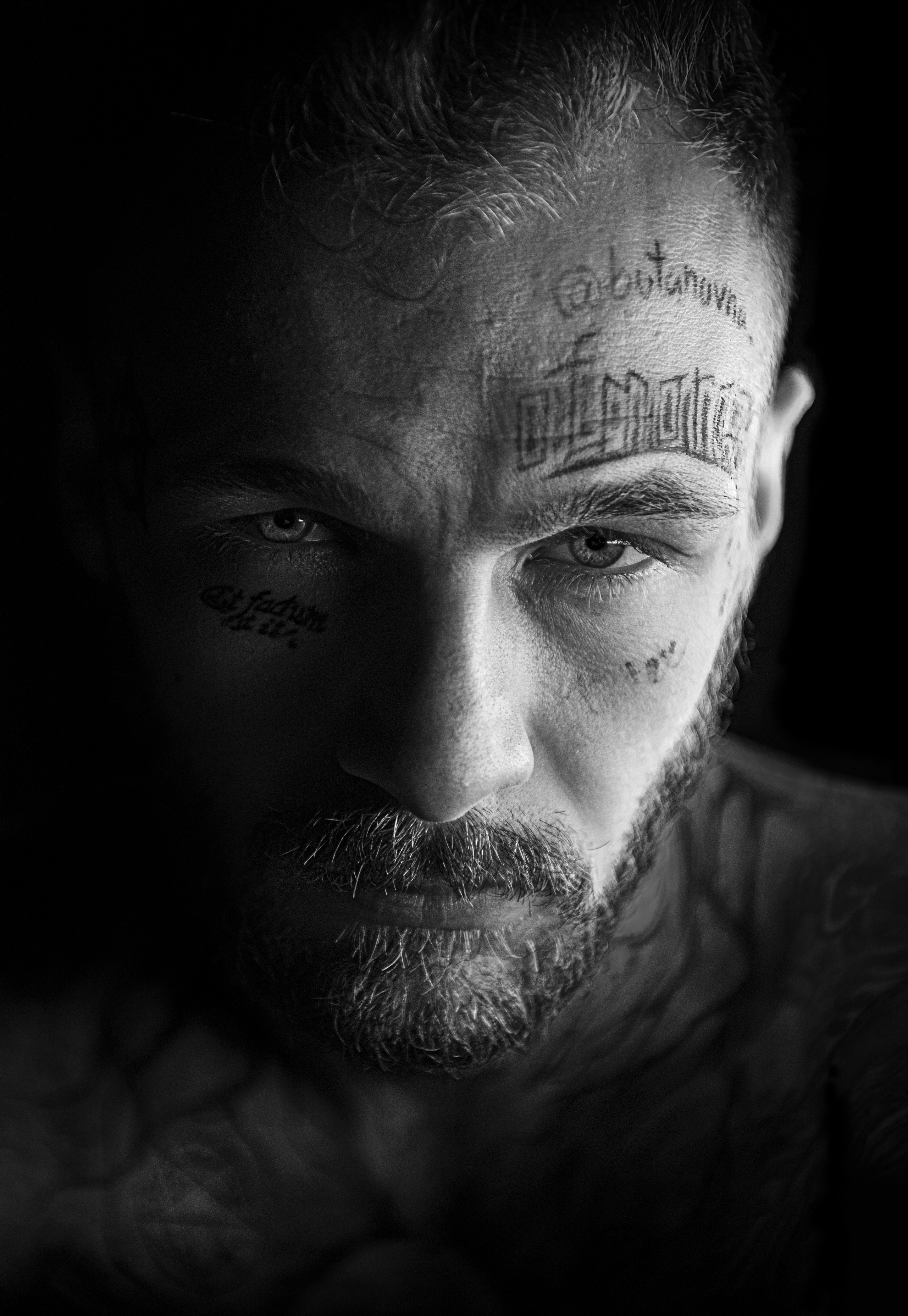 A striking black-and-white close-up portrait of a man with intense eyes and facial tattoos, partially illuminated by soft light against a dark background. The raw emotion and intricate details of the tattoos create a captivating and edgy aesthetic. This image highlights the photographer’s expertise in portrait photography, ideal for clients seeking bold, impactful visuals for editorial, branding, or creative projects.