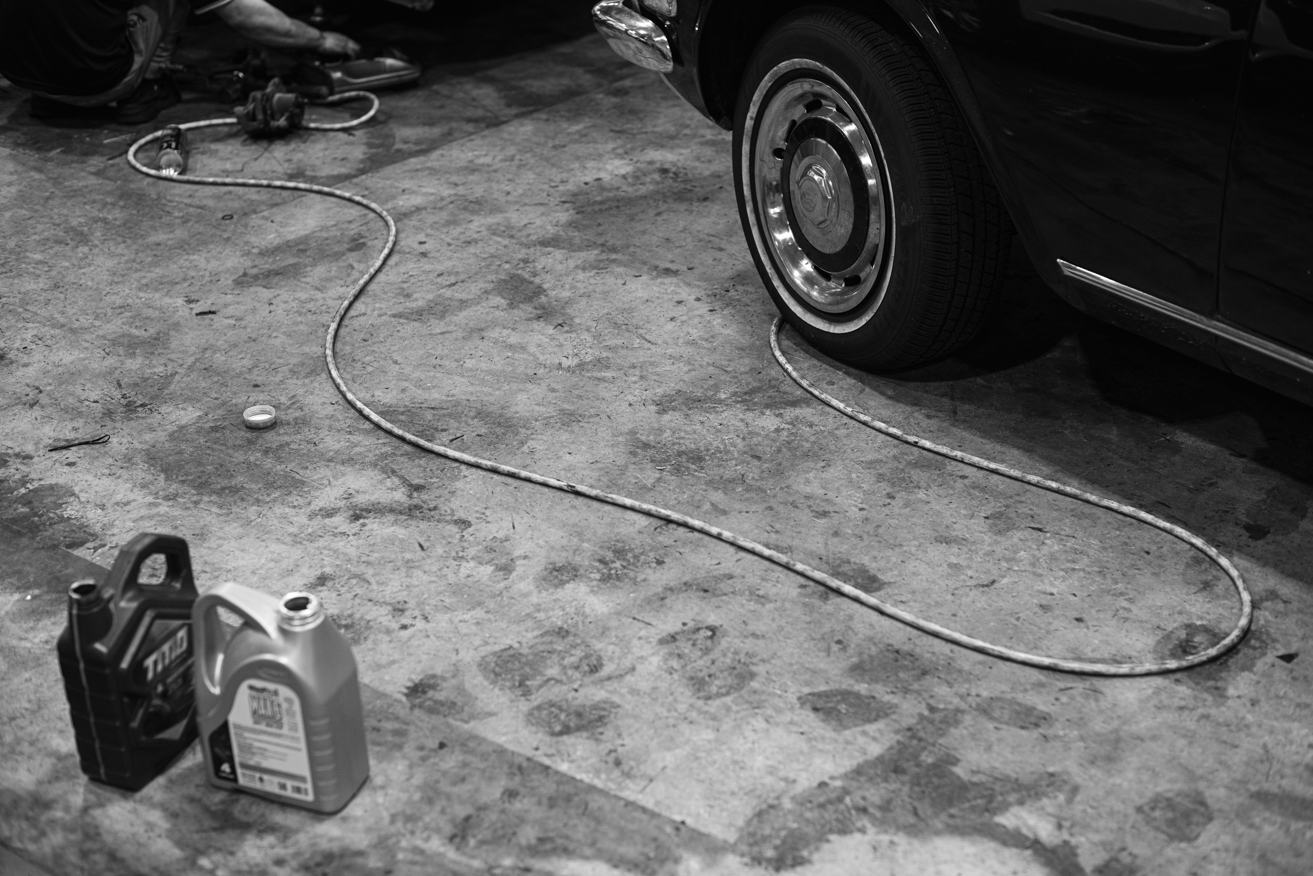 High-contrast black-and-white photo of a classic car wheel, tools, oil containers, and a coiled power cord scattered on the workshop floor at The Garage by Classic Motorworks in Singapore. Captured by MirrorKlezz Photography, this image highlights the gritty details of vintage automotive maintenance, emphasizing the raw craftsmanship involved in classic car restoration. https://www.classicmotorworks.com.sg/