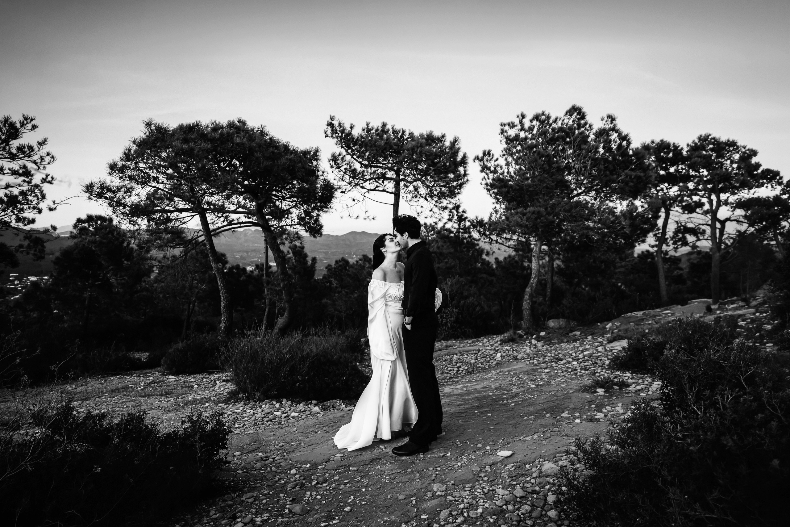 Couple standing together on a rocky viewpoint surrounded by trees during a private mountain elopement in Barcelona, Spain. This black and white destination wedding portrait highlights intimacy and timeless elegance.