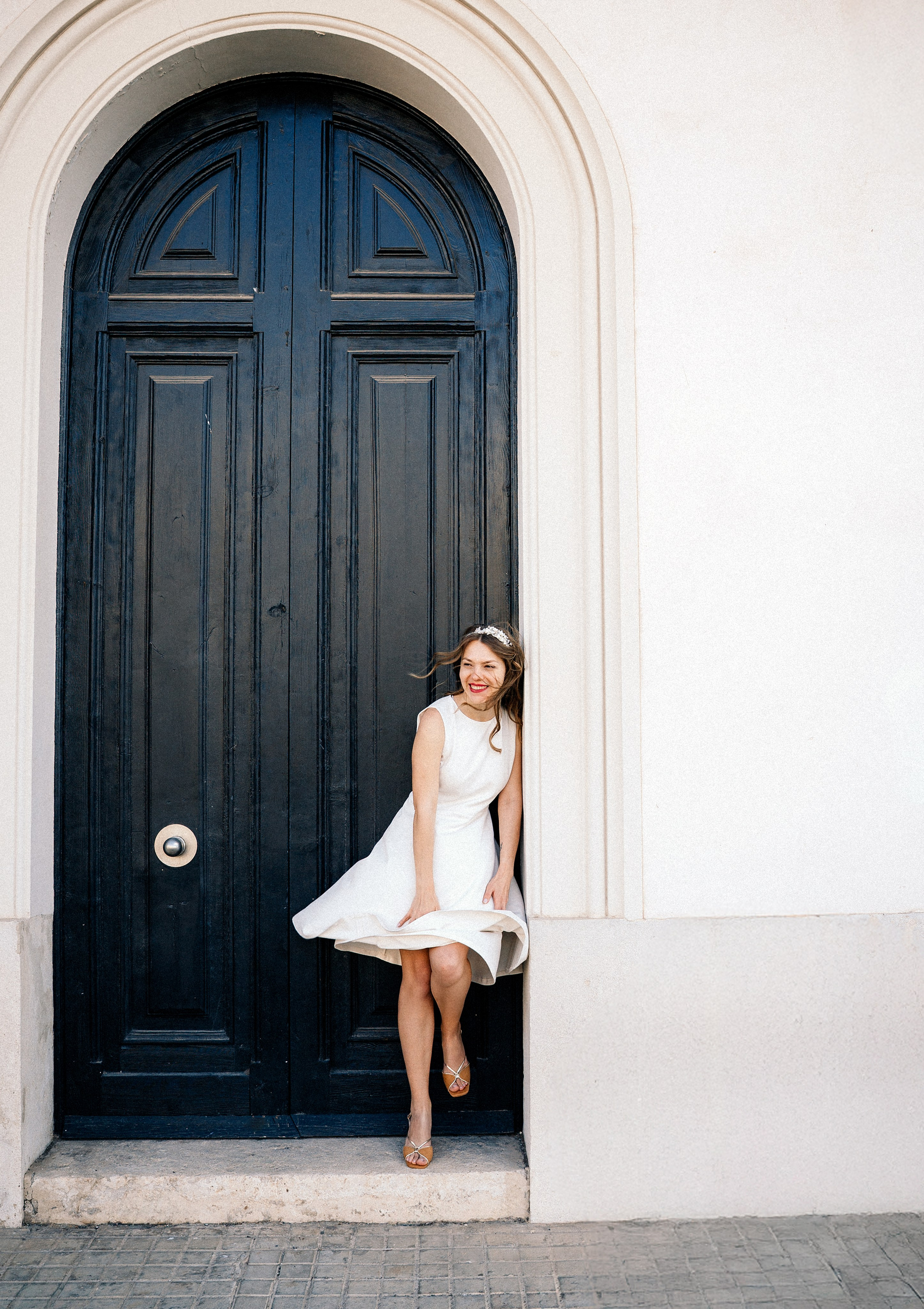 Bride in a short white dress poses gracefully in front of a classic blue door in the historic center of València. This elegant portrait reflects the charm of an intimate city hall wedding in Spain.