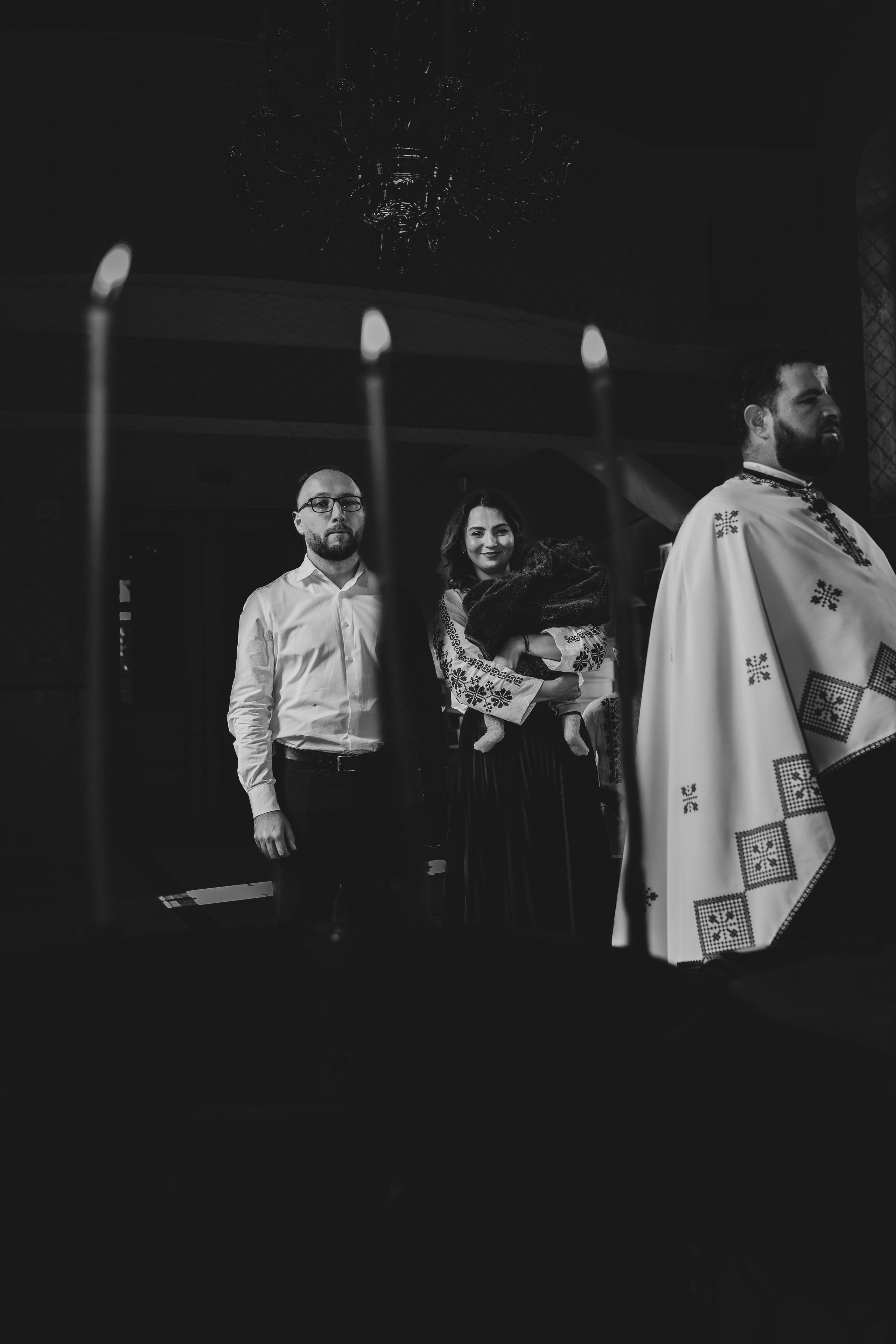 Fotografie artistică alb-negru și minimalistă de la botezul lui Arthur Mihail în Catedrala Bârlad. În centrul unui cadru încadrat de lumânări, preotul, nașii și pruncul sunt izolați, creând o imagine puternică și solemnă a momentului de dinaintea binecuvântării.