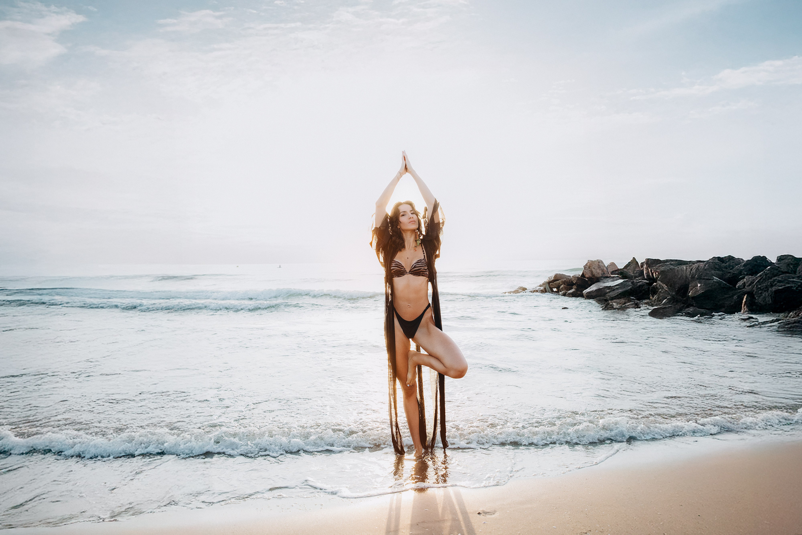 Художественный пляжный портрет на рассвете в Пальма-де-Майорка, Испания — женщина в йога-позе в чёрном бикини и прозрачной накидке. Спокойная и вдохновляющая фотосессия для тех, кто ищет лайфстайл и пляжную съёмку на Майорке и у моря.