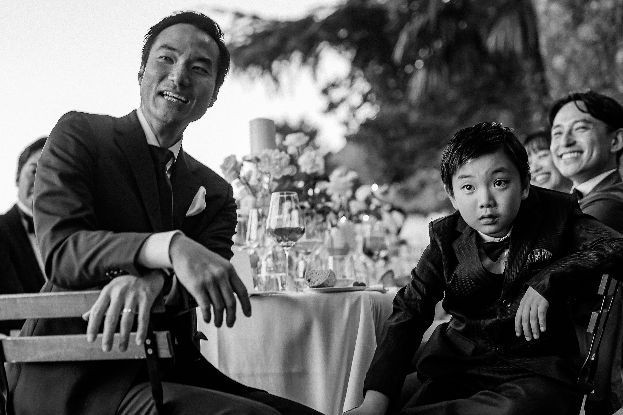 Father and young boy seated at a dinner table, sharing a moment, with lively wedding guests in the background.