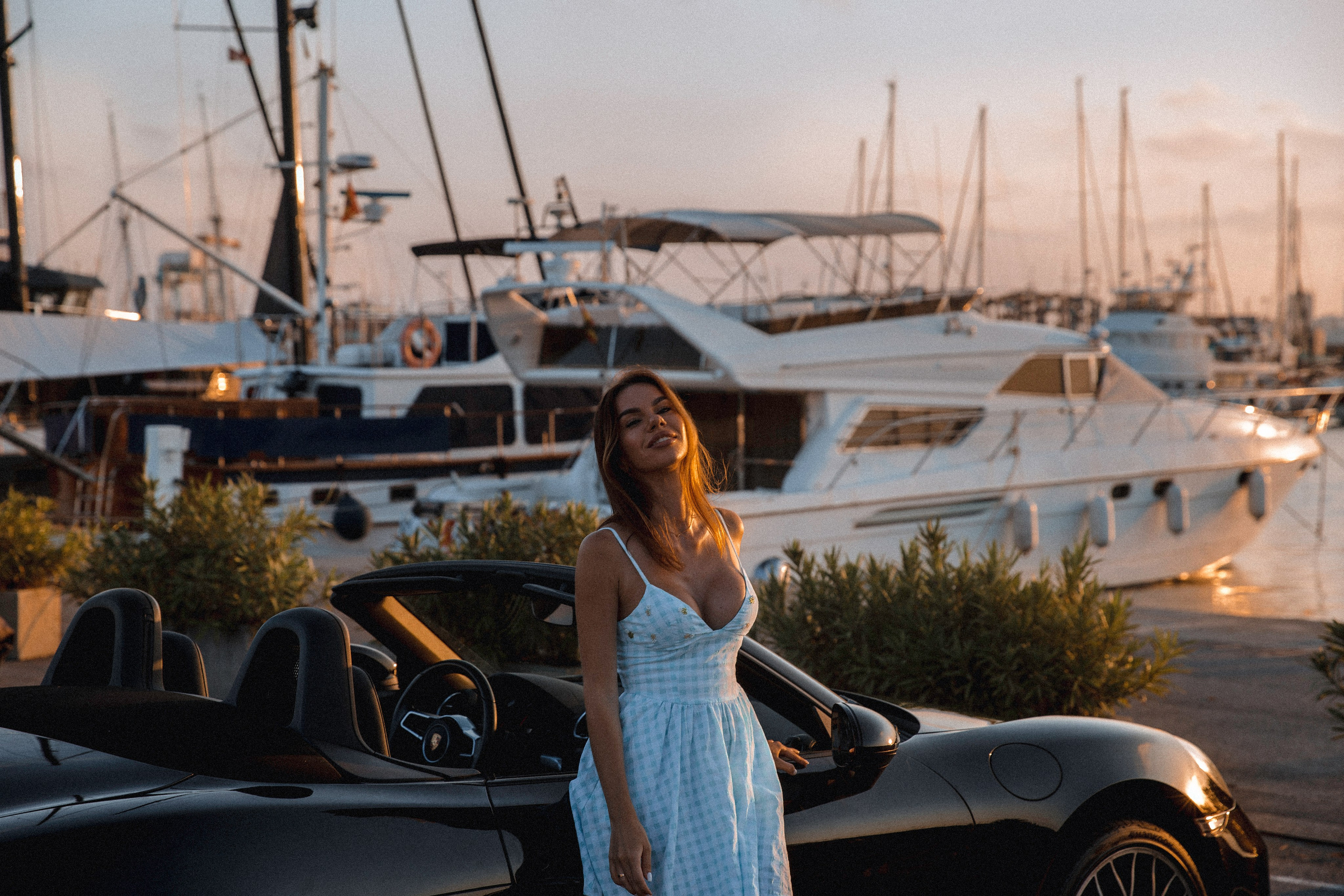 Женская фотосессия в Валенсии, Испания, где уверенная женщина в лёгком летнем платье стоит рядом с кабриолетом у марины в «золотой час», с роскошными яхтами и тёплым средиземноморским светом на фоне — идеально для женских фотосессий в Валенсии, лайфстайл- и fashion-портретов и профессиональной женской фотографии по всей Испании.