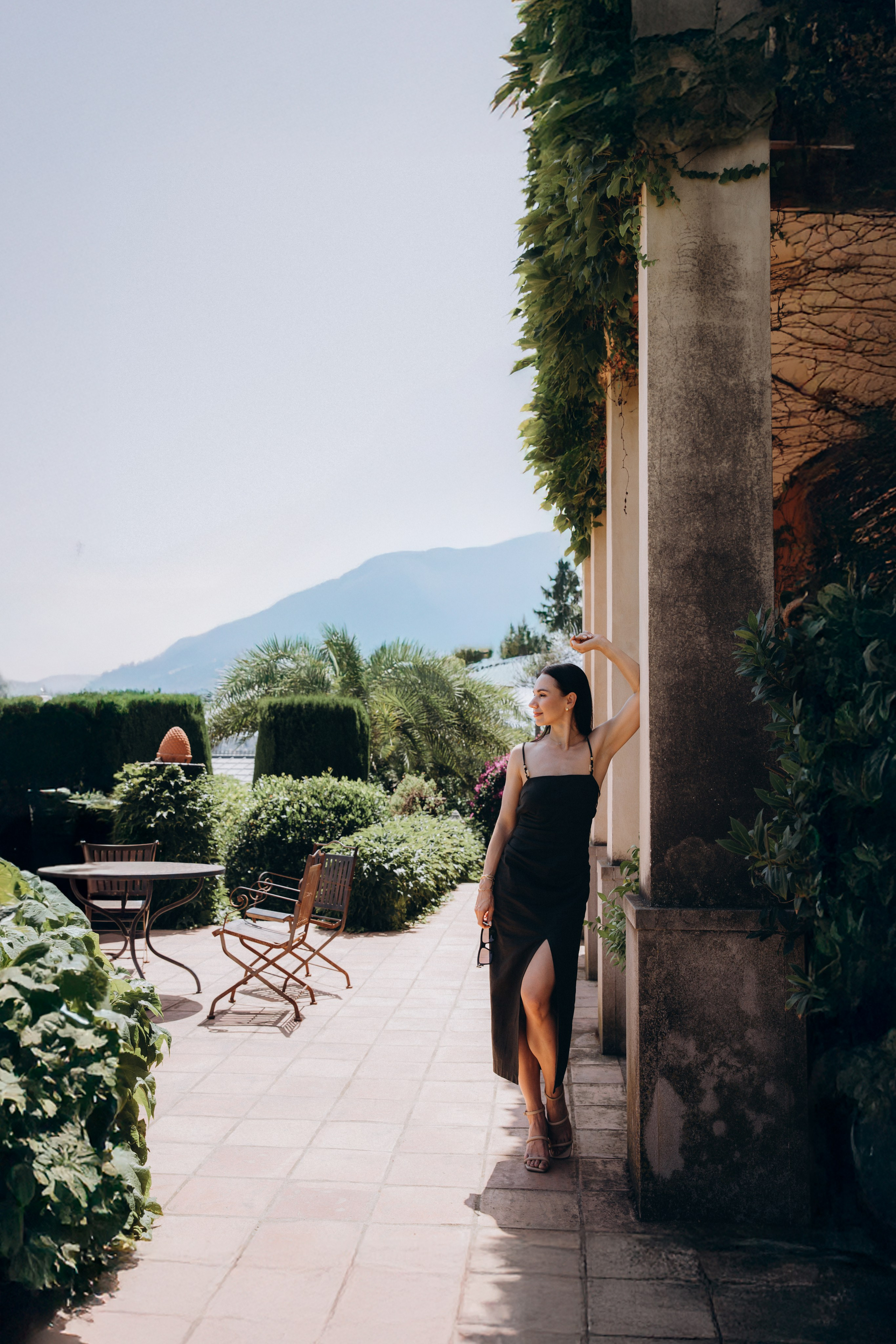 Женщина в черном платье лежит на кованой скамейке в пышном тропическом саду с драматичным светом и архитектурными тенями в Аликанте, Испания — креативная и выразительная портретная фотография, сочетающая элегантность, природу и современный дизайн, идеально подходит для портретных фотосессий в Аликанте и Испании.