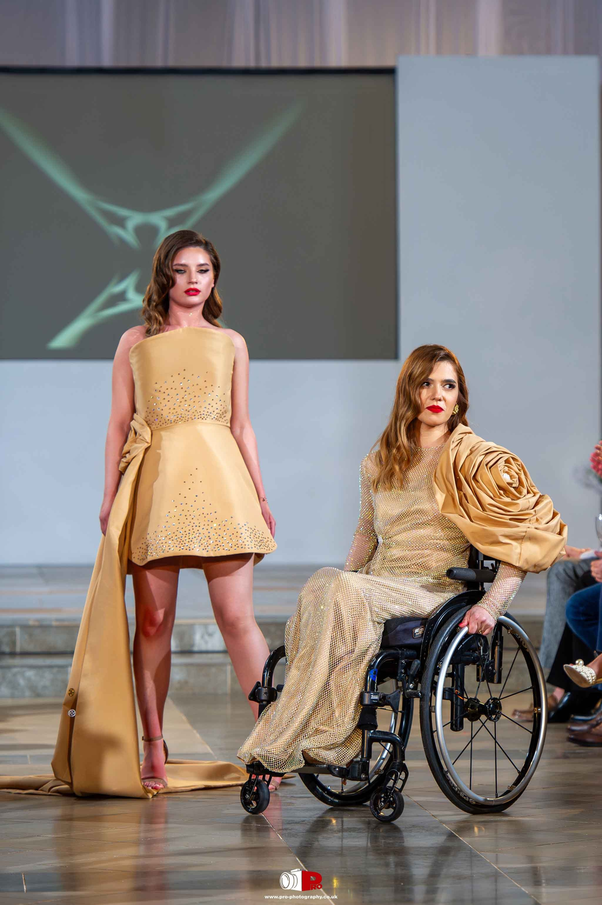 Two models are showcasing glamorous gold dresses on a fashion runway, and one is seated in a wheelchair, representing inclusivity in fashion.