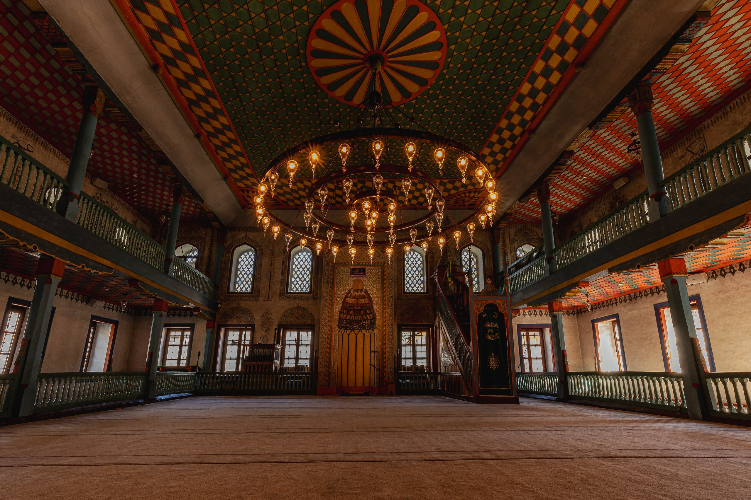 Interior architectural photography of a historic Ottoman mosque featuring geometric ceiling patterns, arched windows, wooden galleries and decorative chandeliers illuminated by natural light.