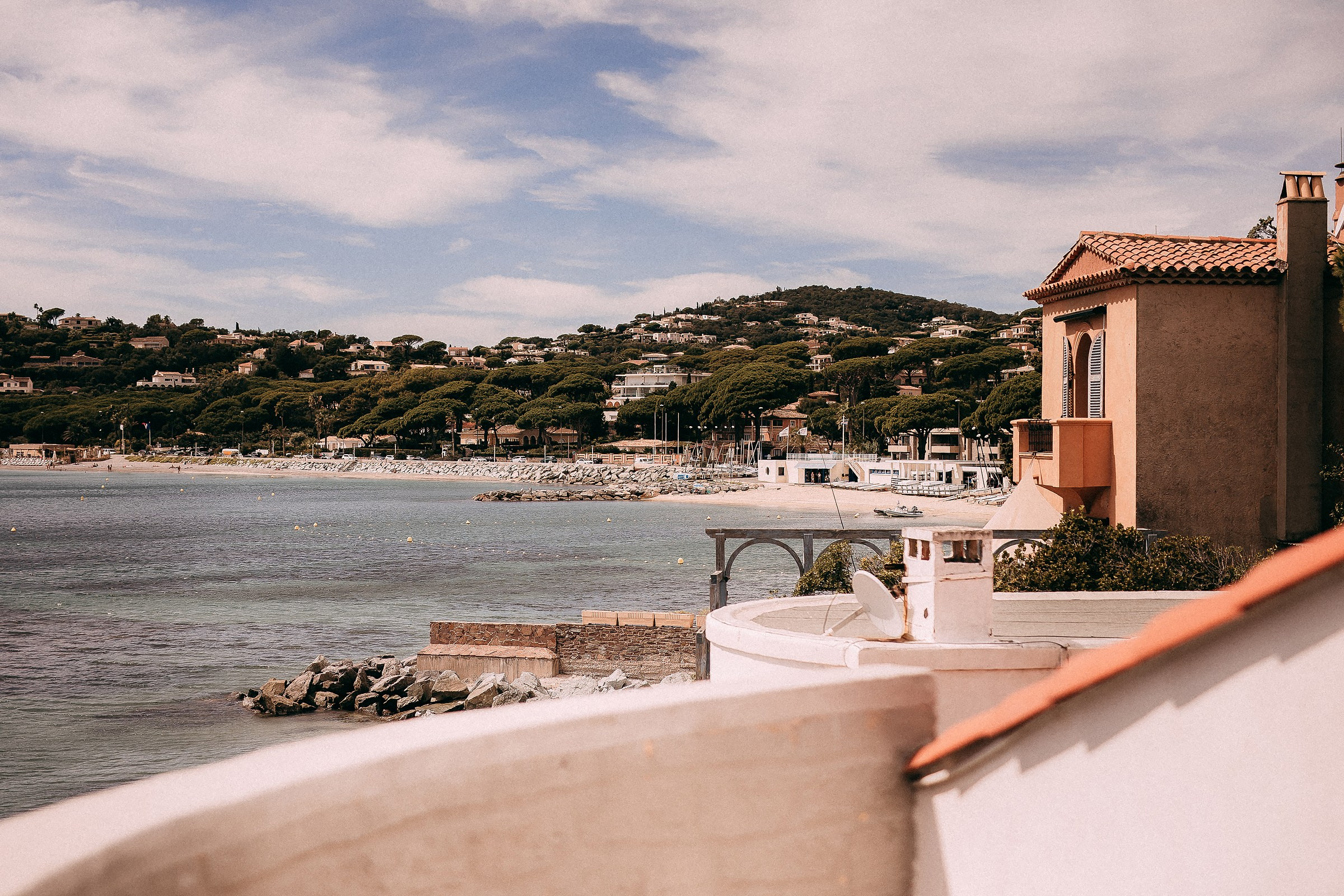 A serene view of a calm coastline in Provence, France, with a charming Mediterranean-style house overlooking the clear water and lush hills in the distance.