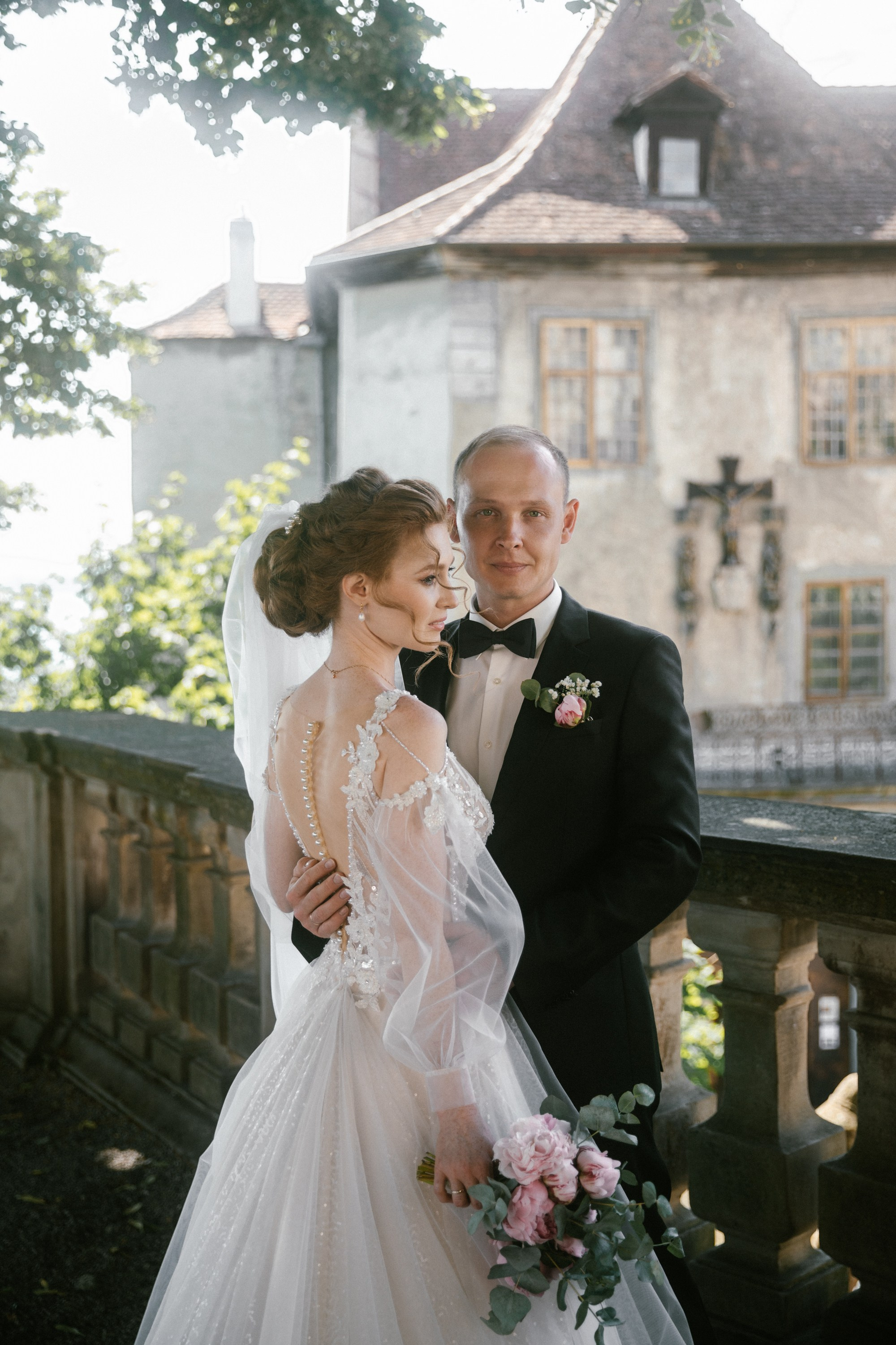 Bride and groom posing on shaded terrace at Schloss Meersburg, bouquet of pink flowers visible