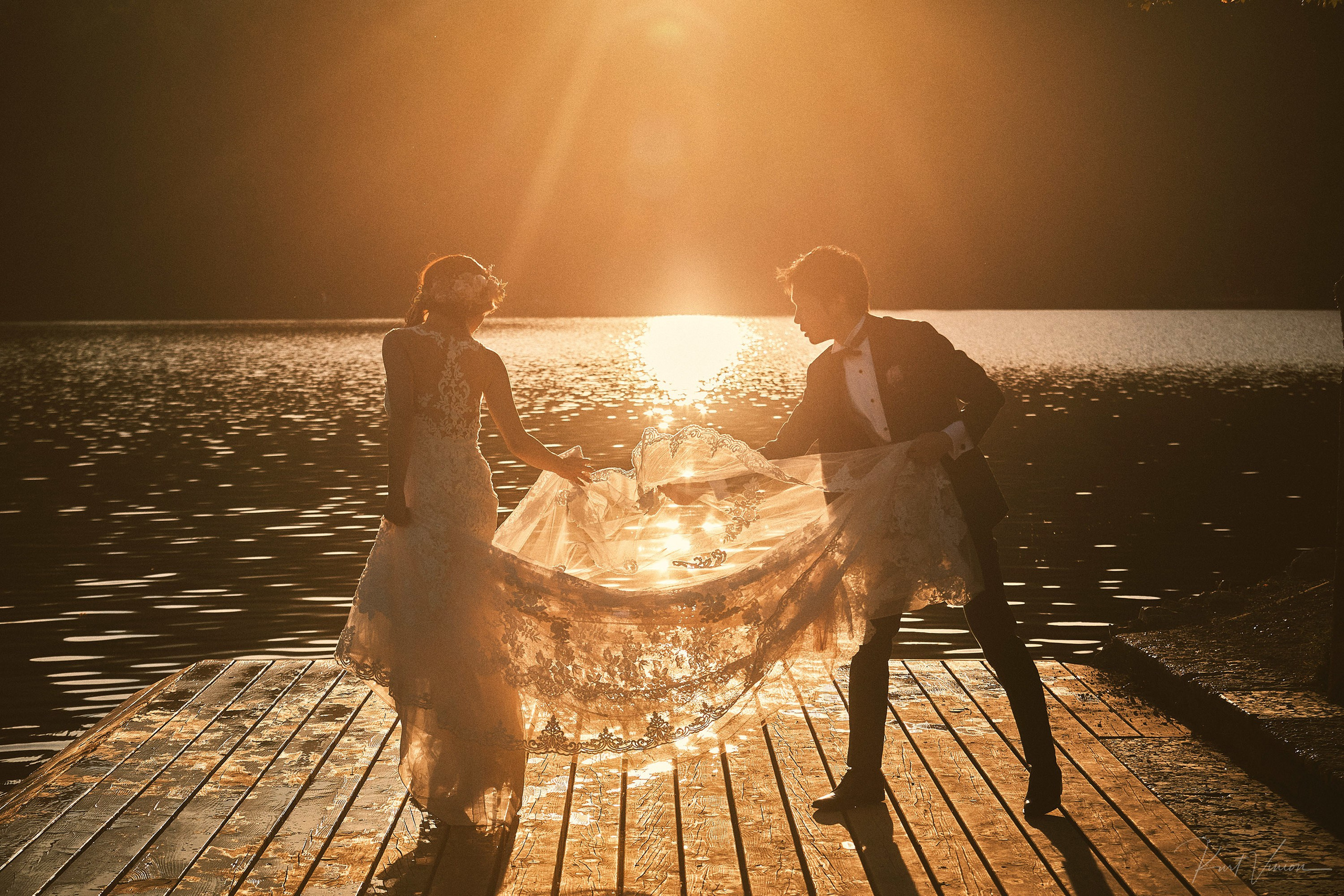 Groom tending bride's gown near-silhouette golden sunset flare