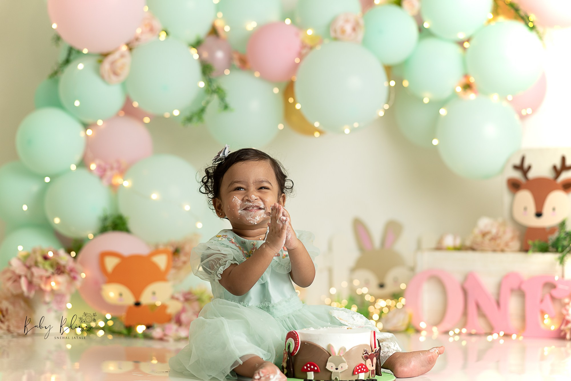 one year old girl smashing the cake with mint green balloon arch setup in the background.