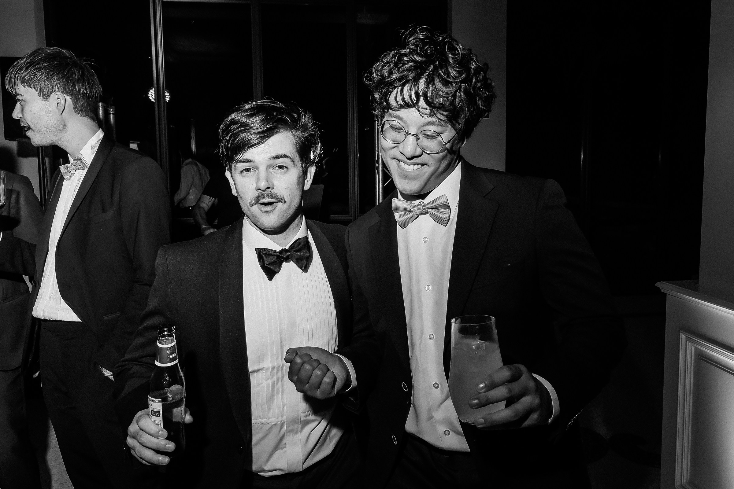 Two men in tuxedos, one with curly hair and glasses, smiling and holding drinks during a lively indoor gathering.
