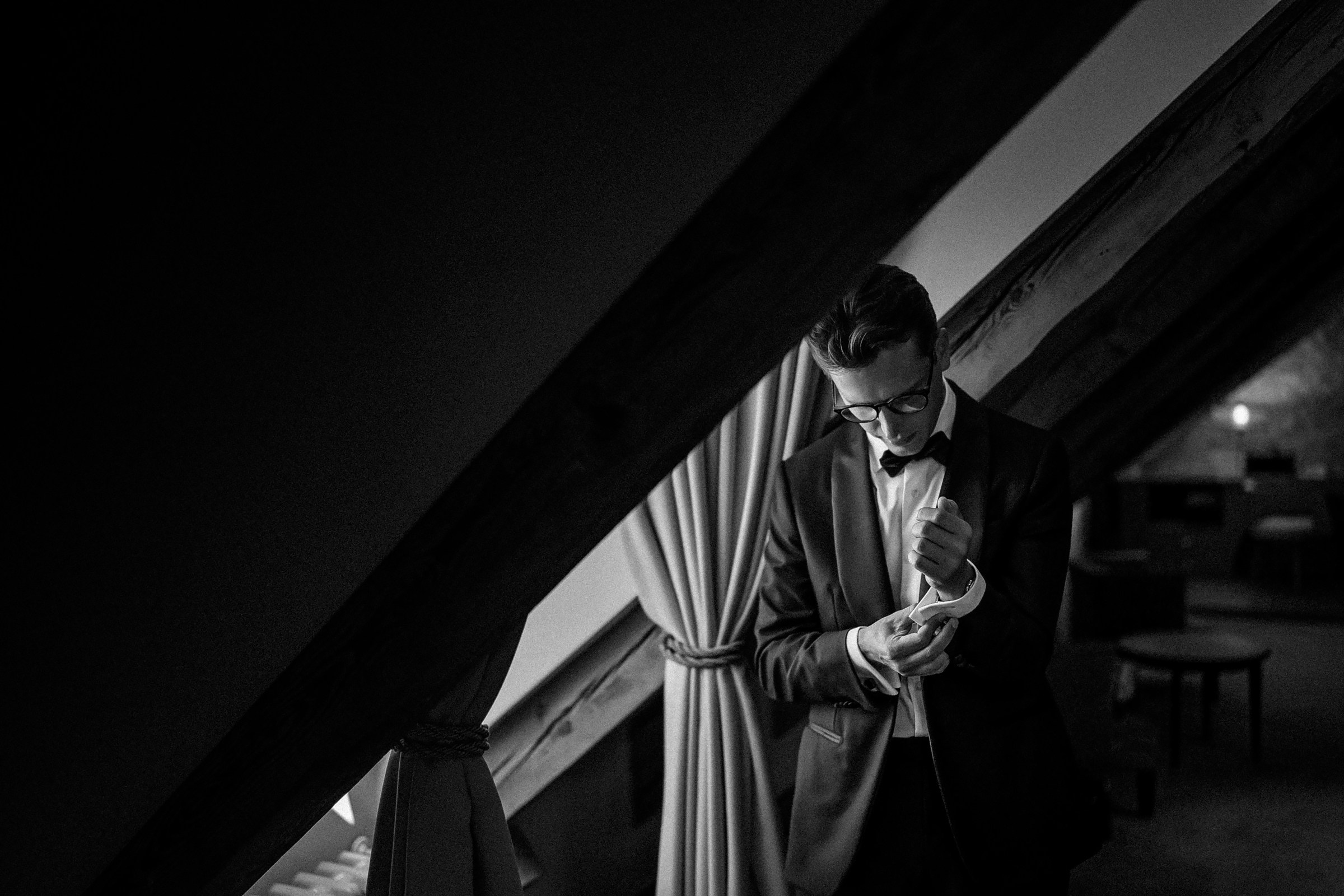 The groom in a suit is adjusting the cuff of a shirt while standing in a room with slanted wooden beams and curtains. The image is in black and white, creating a dramatic and formal atmosphere.