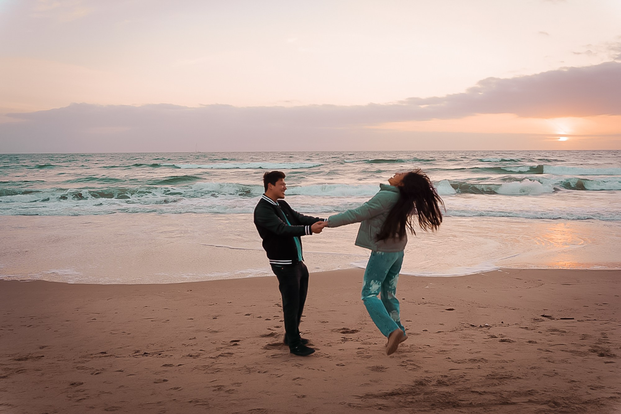 Just-engaged couple spinning on the ocean shore, surrounded by the ocean breeze and sunset light