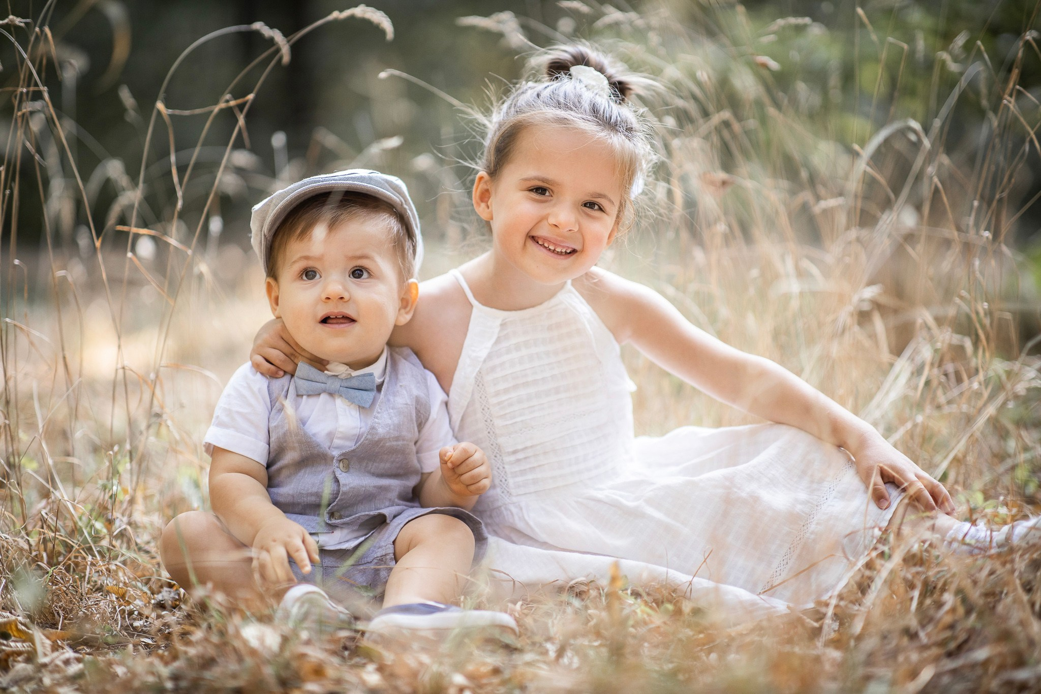 Srećni brat i sestra sede na sunčanom polju. Slatka beba dečak u šeširu i devojčica u beloj haljini, porodični portret na otvorenom.