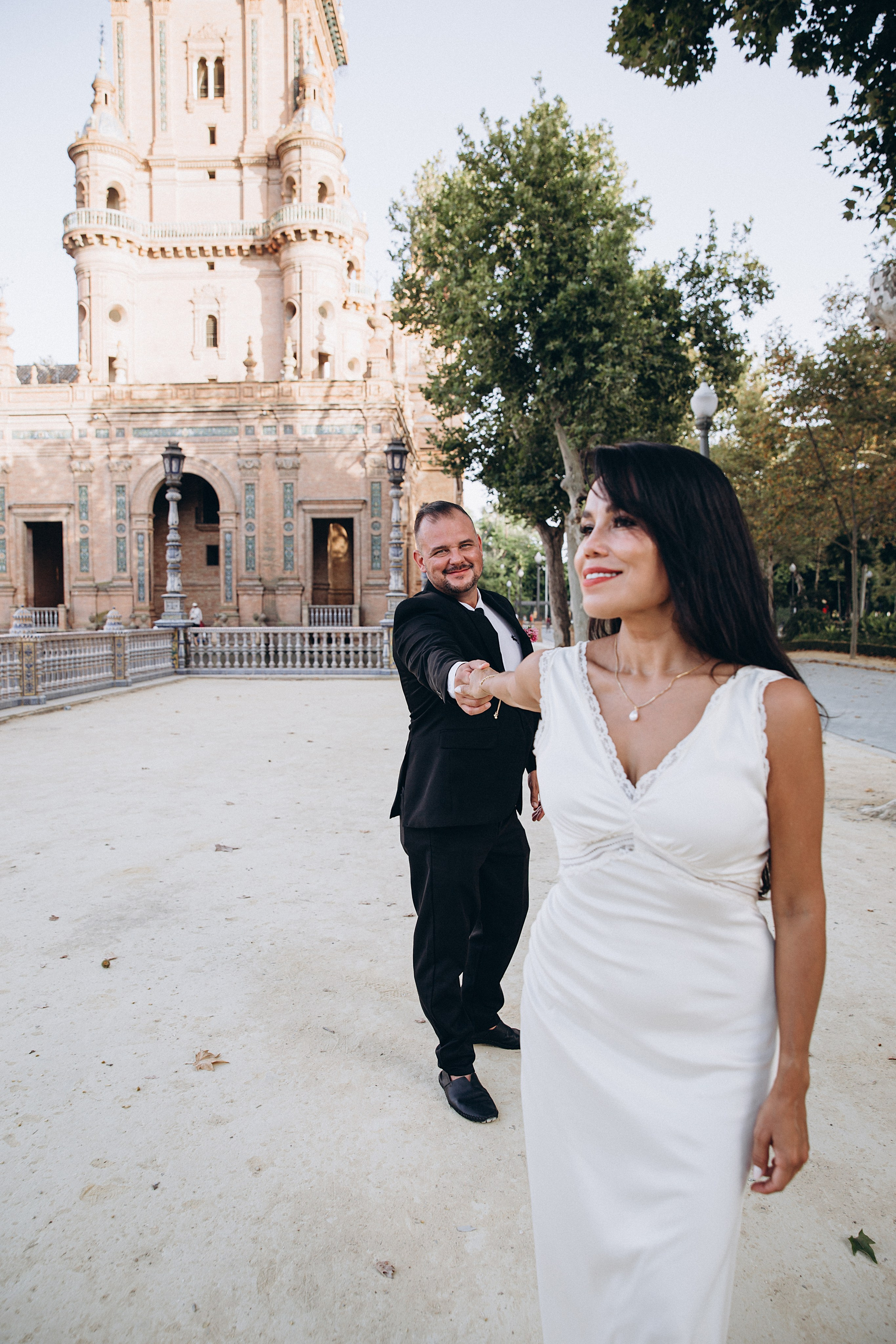 Bride walking ahead while the groom follows, reaching for her hand in Plaza de España, Sevilla. Playful documentary-style moment from their civil wedding celebration in Andalusia.