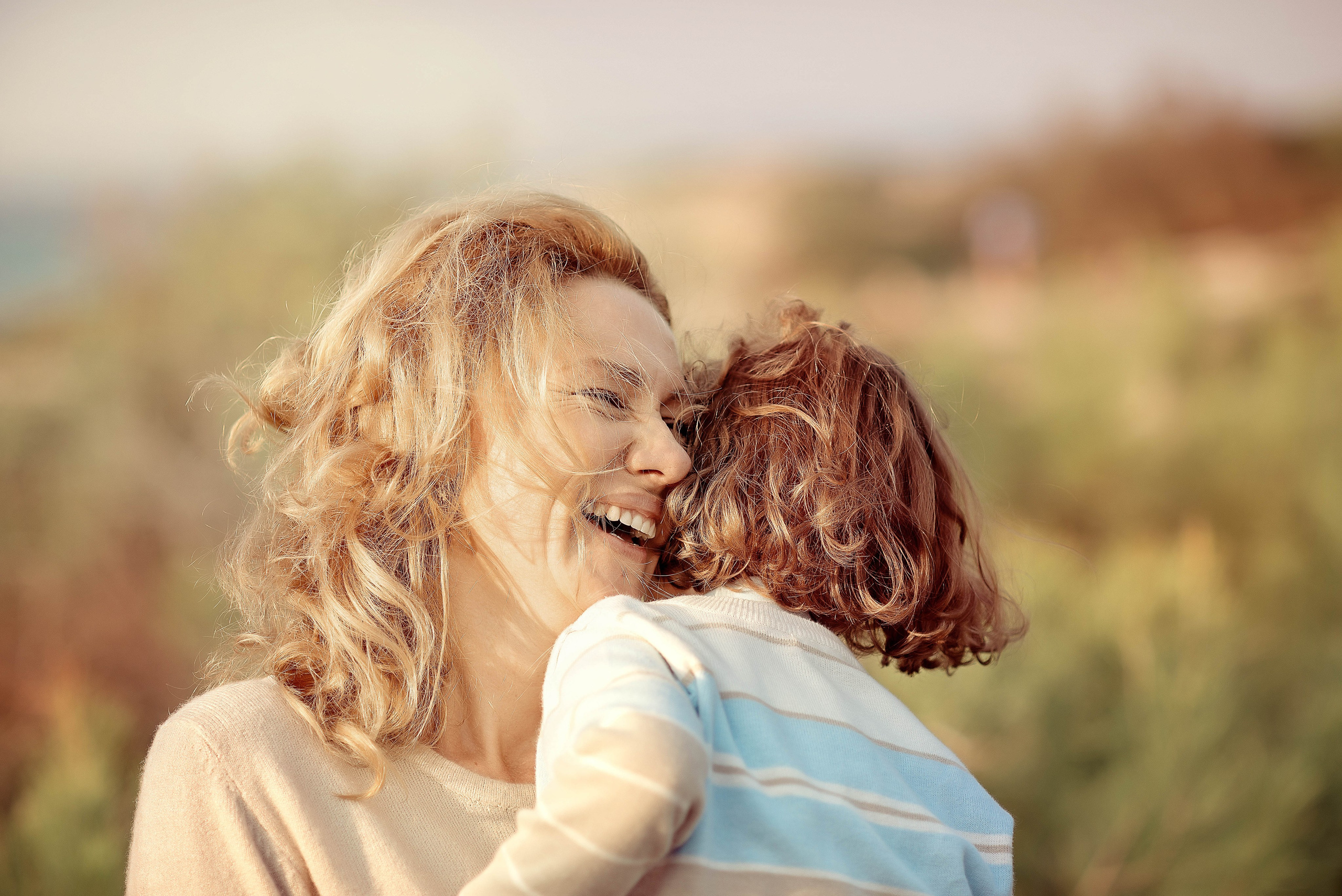 A mother smiles joyfully while holding her curly-haired child in her arms during golden hour, surrounded by soft greenery.