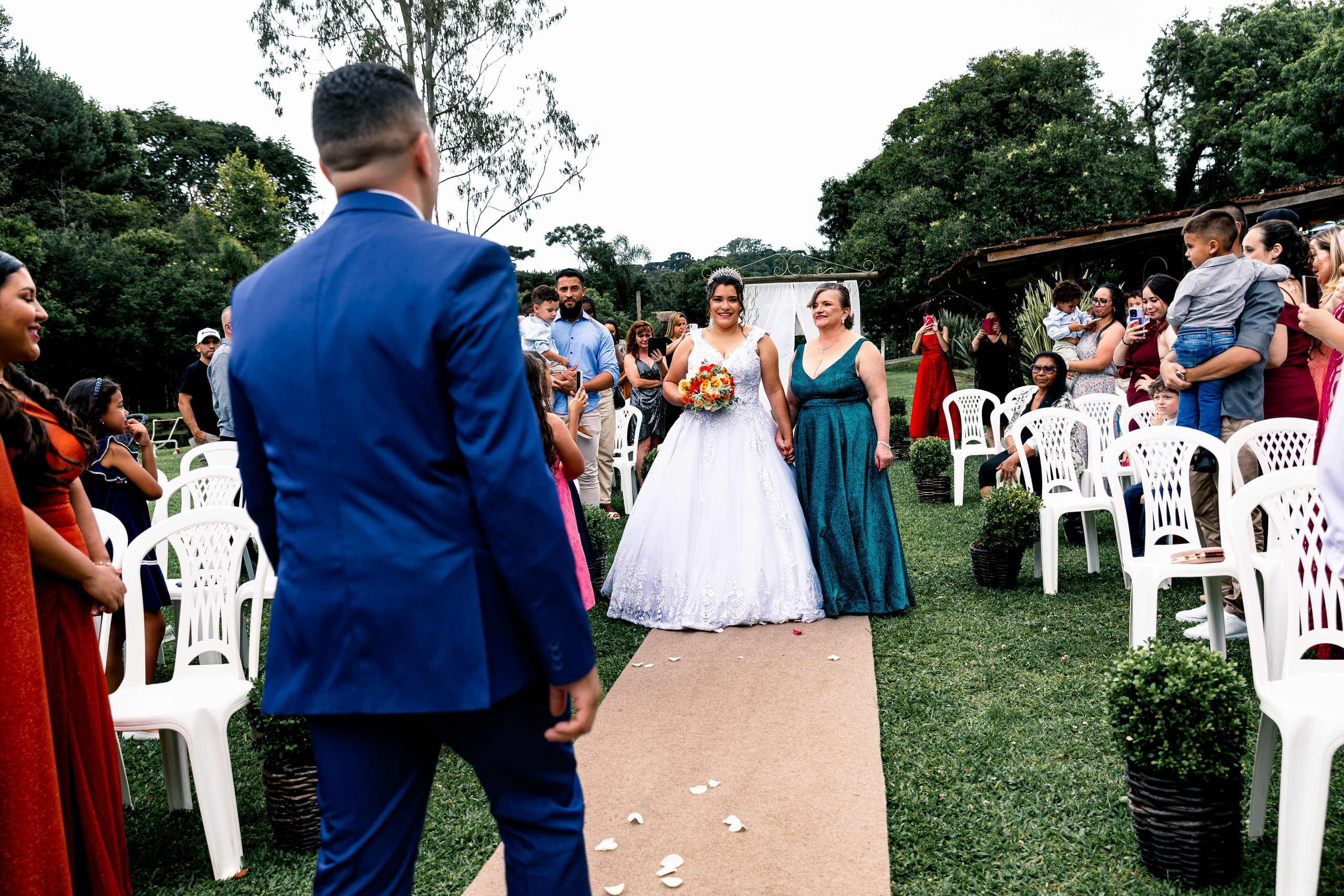 Noiva e mãe caminhando lado a lado no corredor da cerimônia ao ar livre, entre olhares e registros. Fotografia de casamento em Curitiba.