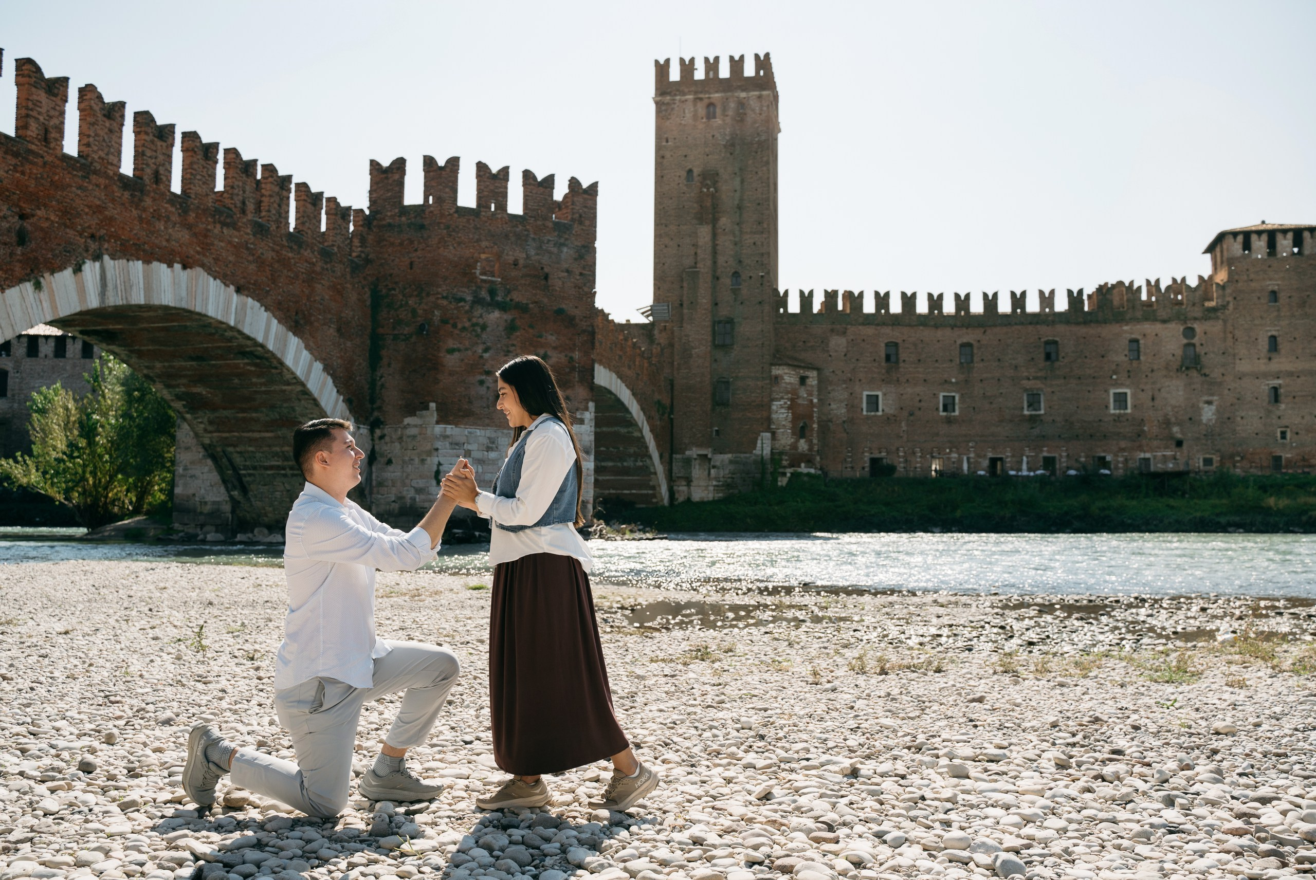 One partner kneeling to propose by the Adige River in Verona, overlooking Castelvecchio.
