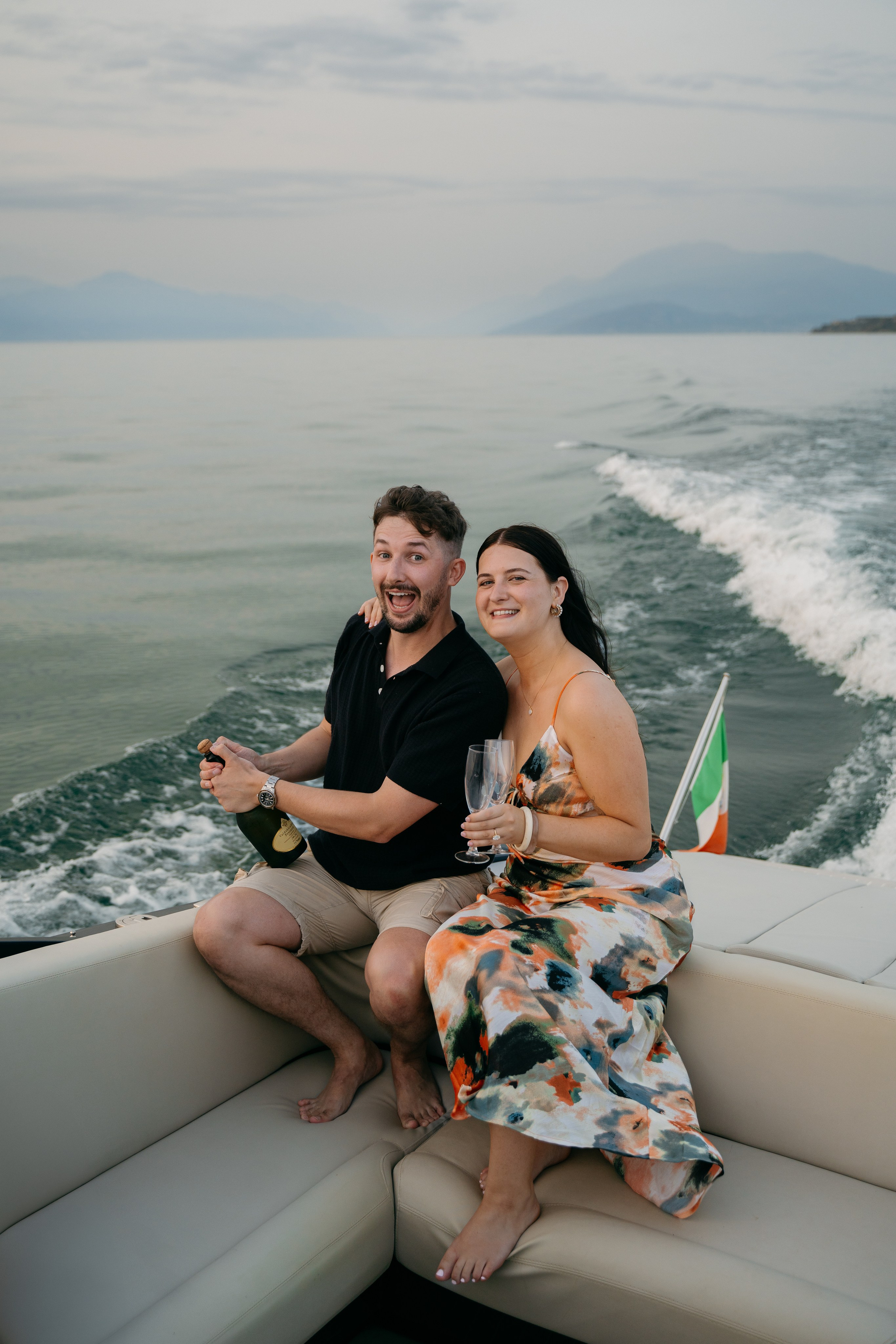Joyful newly engaged couple opening champagne surrounded by golden reflections on a private Lake Garda cruise.