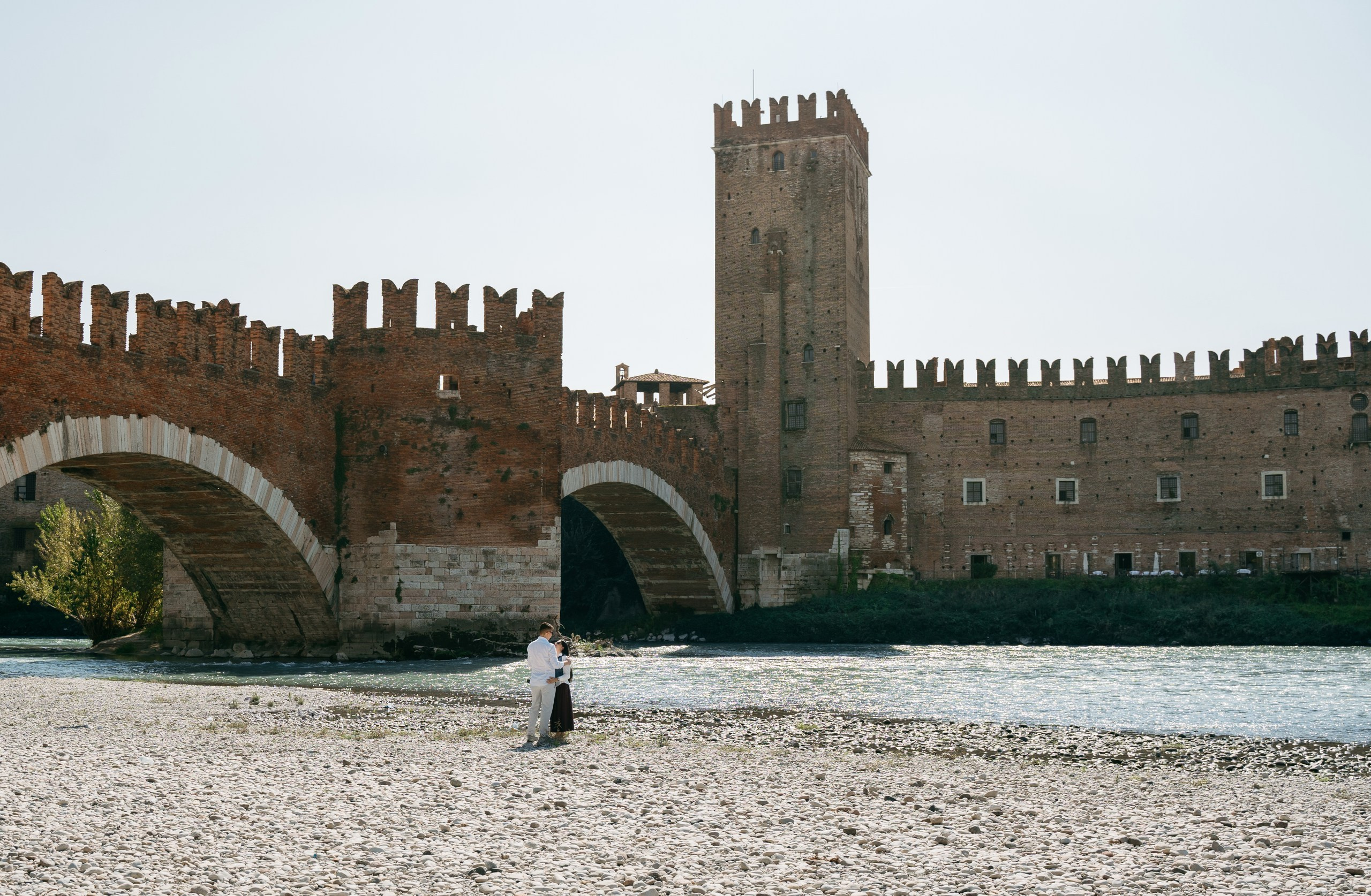 A wide view of a marriage proposal by the Adige River, with Verona’s Castelvecchio behind the couple.