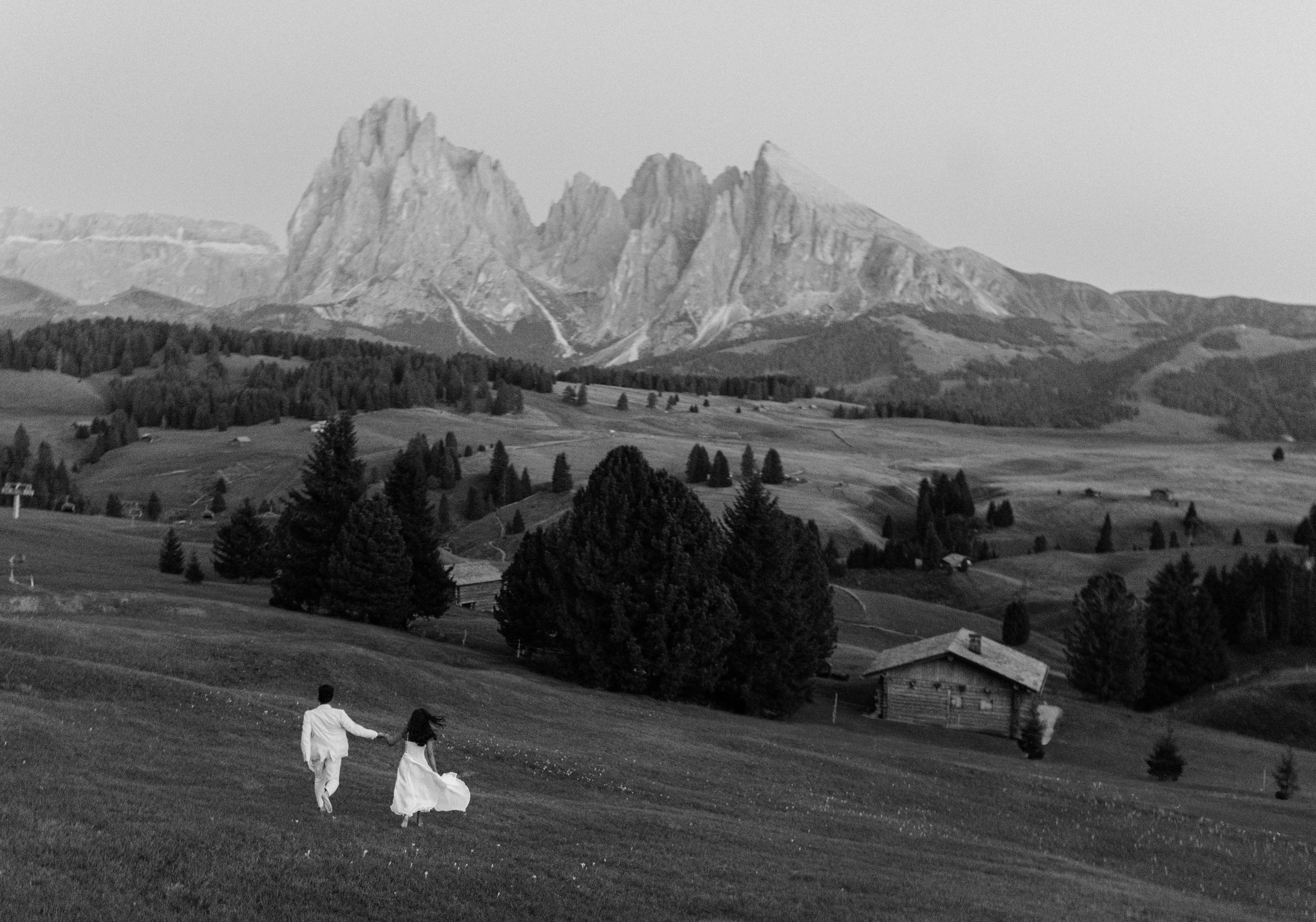 Newly engaged couple running hand in hand towards the mountains on Alpe di Siusi.