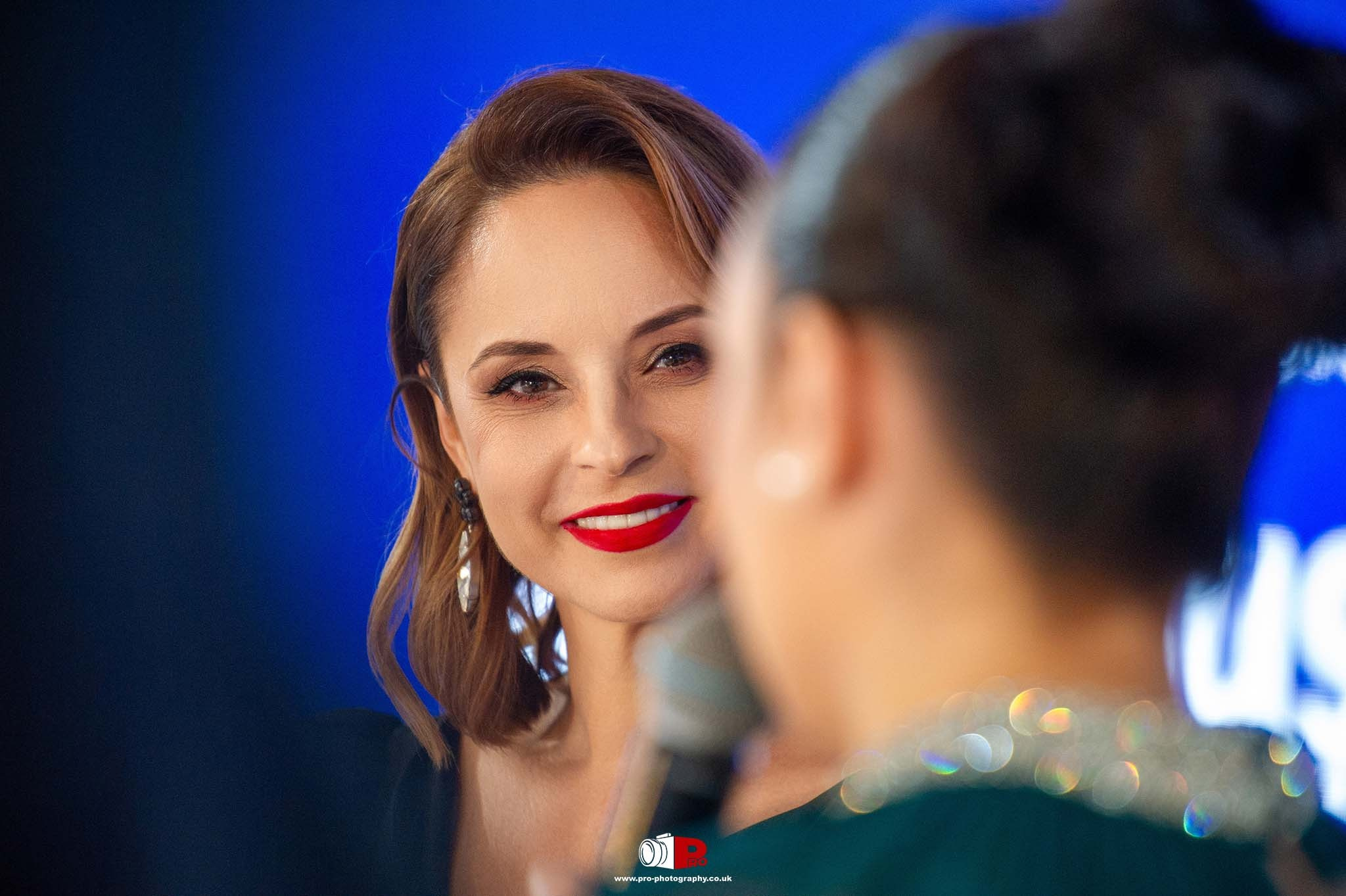 Andreea Marin, with bold red lipstick, is smiling while listening to a speaker at a gala event.