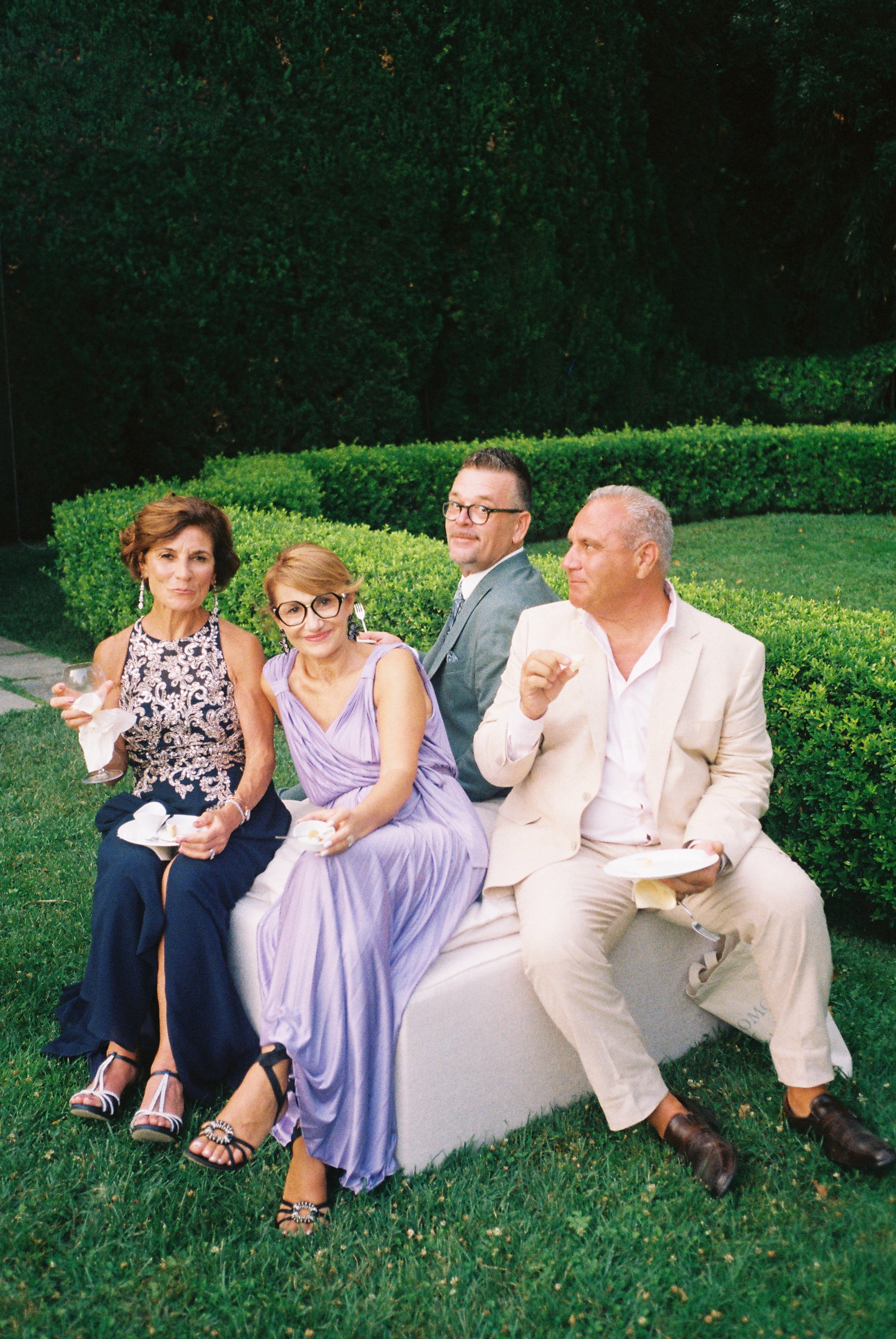 Group of elegantly dressed people seated outdoors, enjoying drinks and conversation in a garden setting.