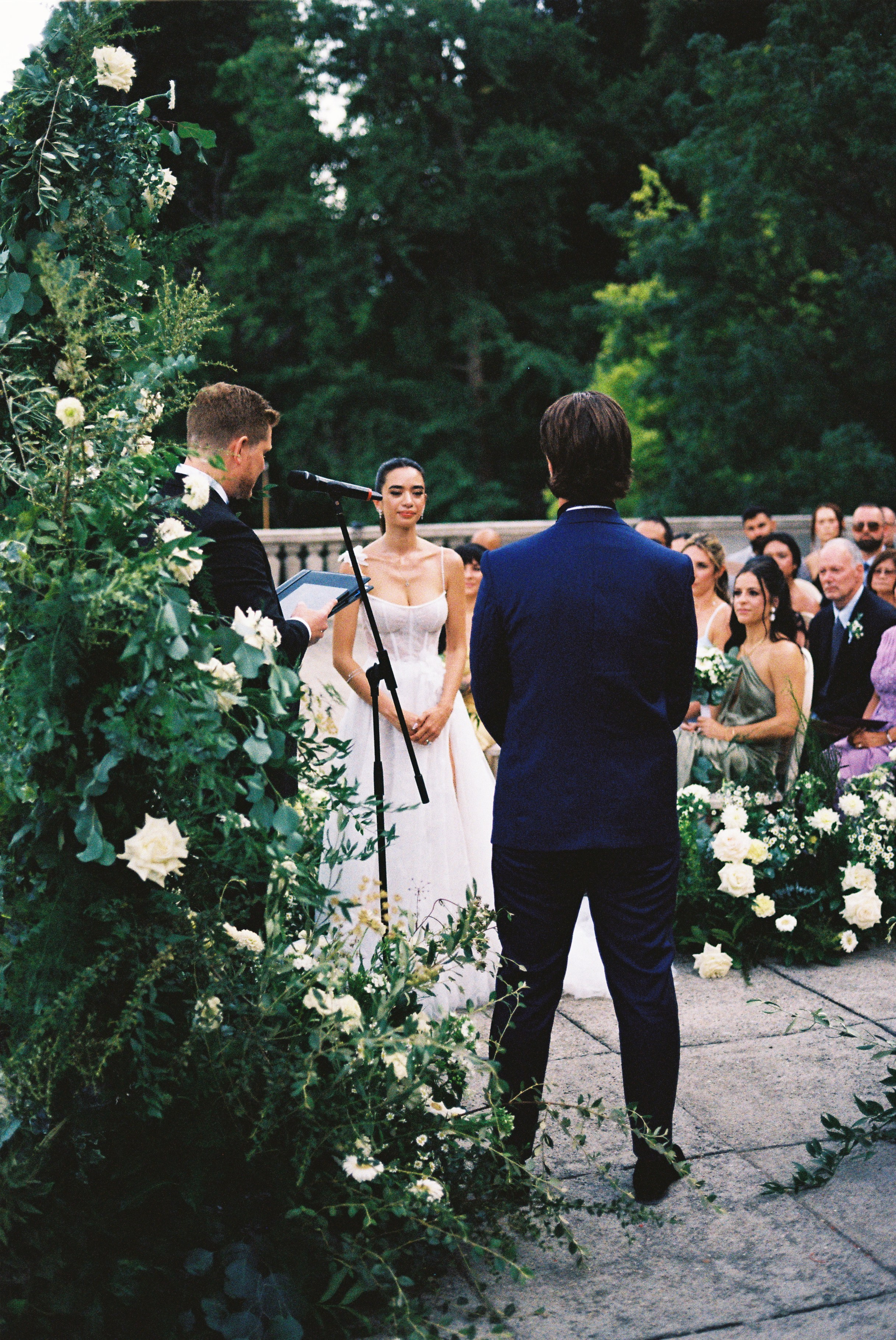 Bride and groom stand together as officiant reads vows, surrounded by lush green floral arch.