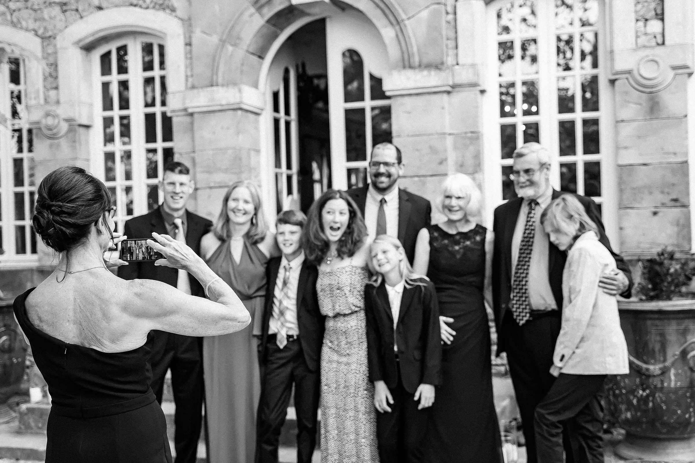 A family portrait being captured in front of a grand chateau in Provence, France, showcasing a mix of formal attire and joyful smiles as a woman photographs the group.