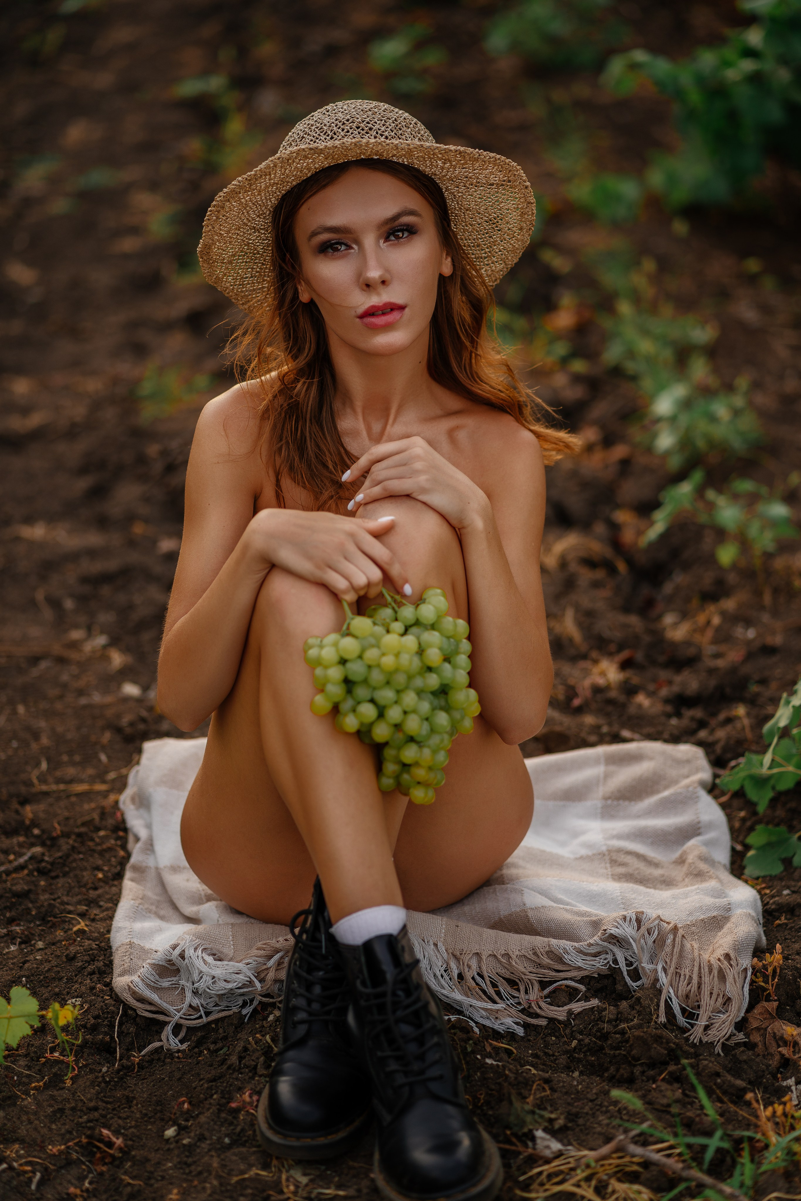 Harvest Goddess. Naked beautiful young woman sits in the vineyard, exuding elegance and tranquility. She holds a bunch of grapes, her body adding a touch of purity against the green backdrop. The serene environment and the model's poise make this image ideal for marketing campaigns or lifestyle promotions.