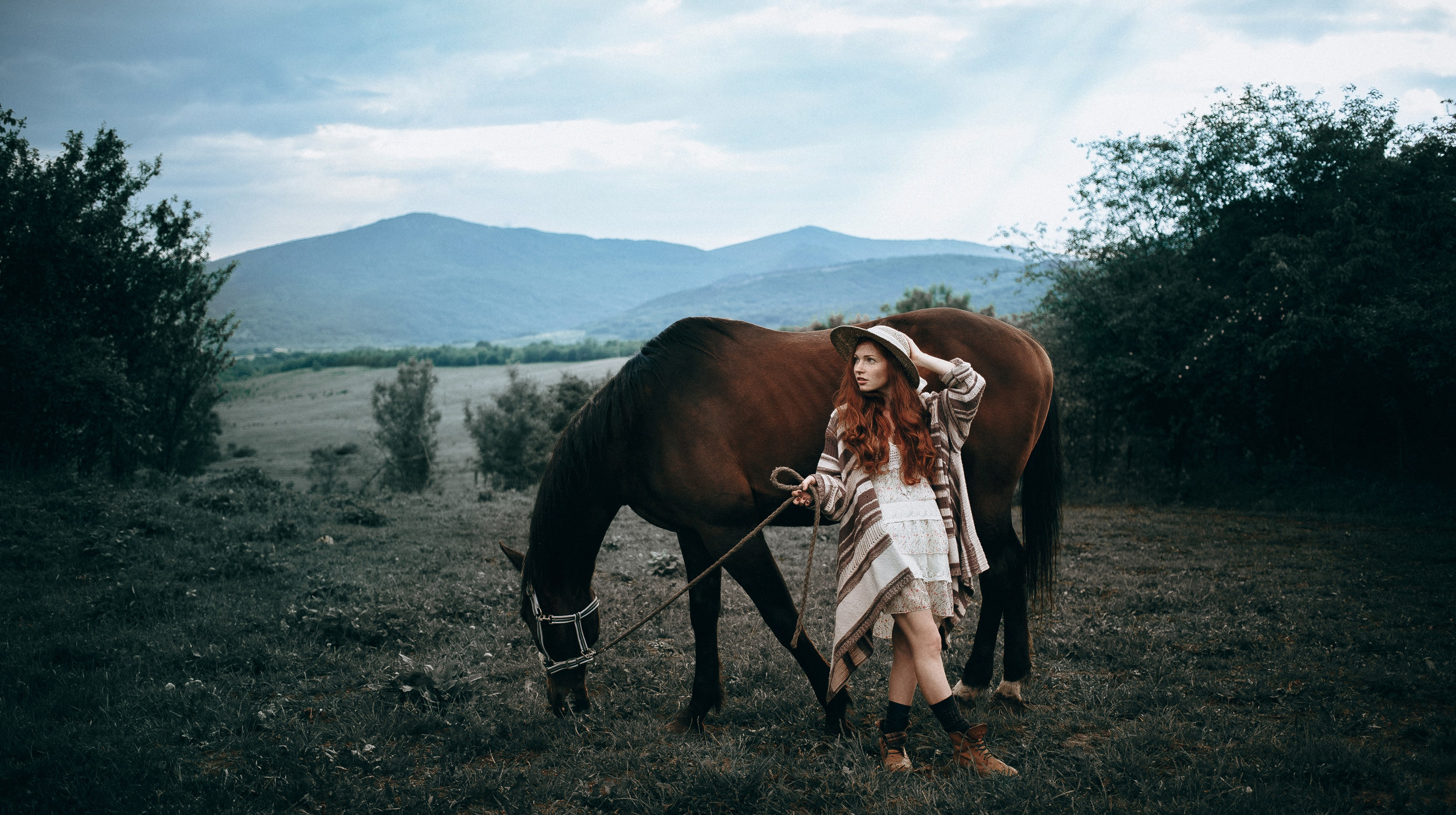 A woman stands next to a horse against the backdrop of majestic mountains. The horse looks powerful and calm, and the model looks confident and peaceful. The mountains in the background create an impressive and inspiring landscape, emphasizing the strength and grace of the horse, as well as the connection between man and nature