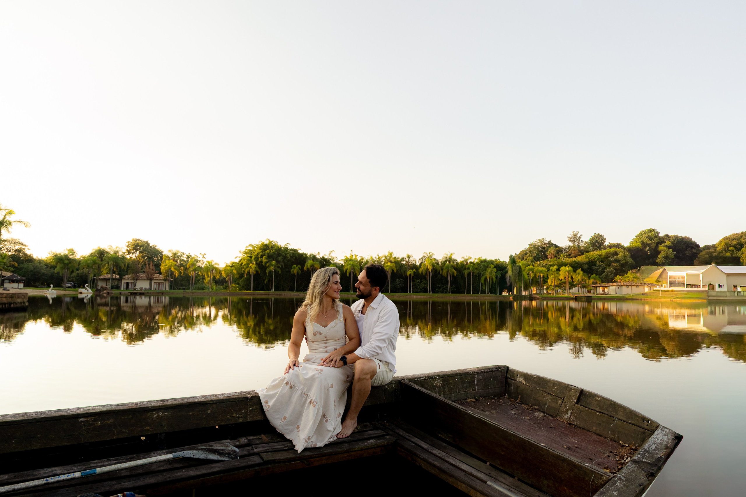 Casal sentado na borda de um barco de madeira se olhando com o pôr do sol dourado e o lago do Resort das Oliveiras ao fundo