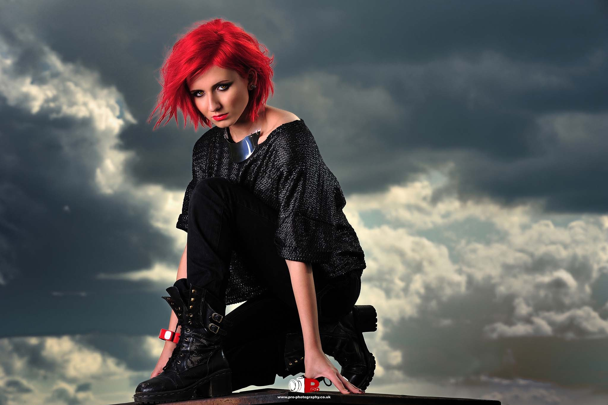 A striking red-haired woman in black fashion crouching under a dramatic cloudy sky.