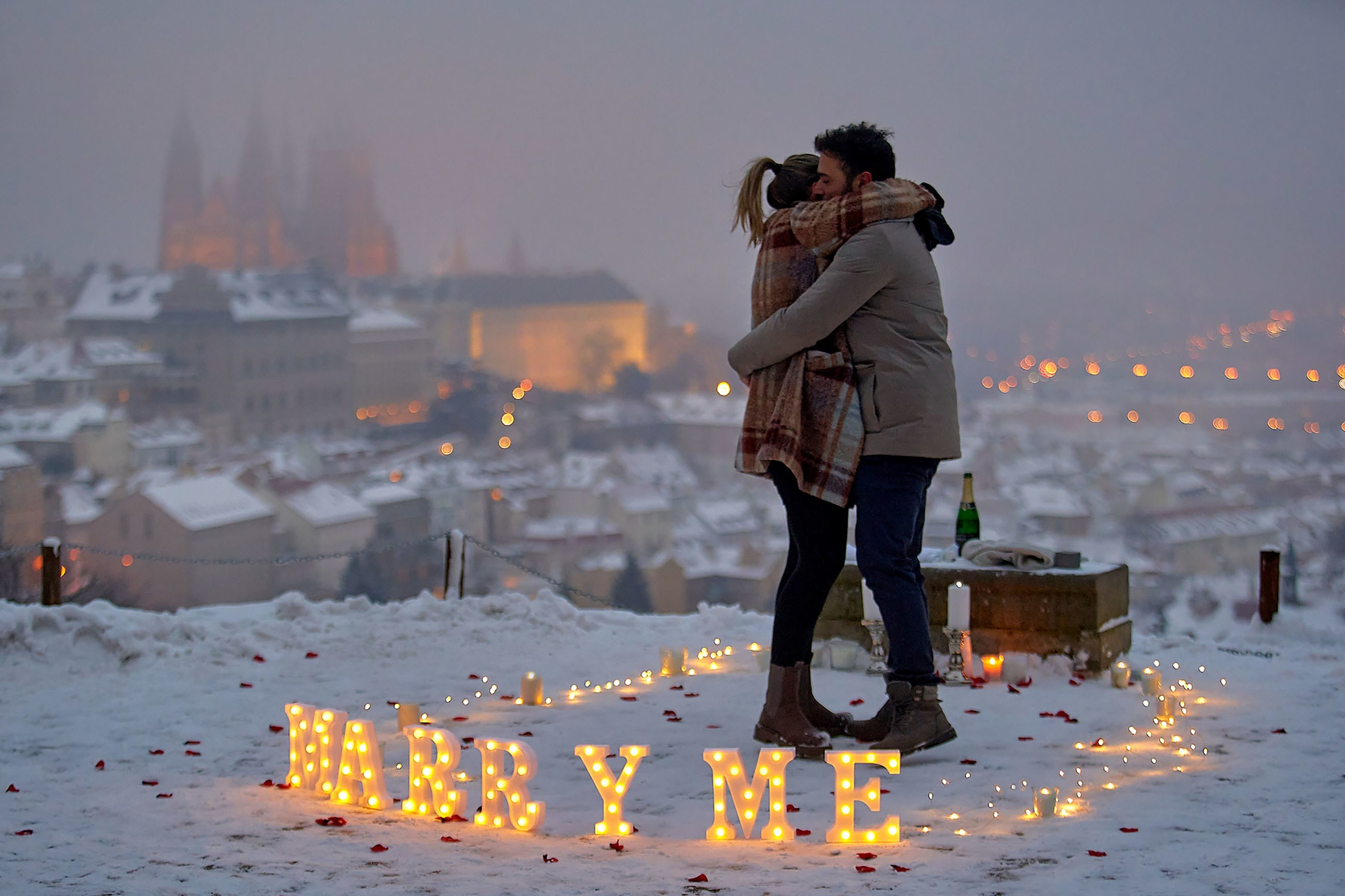 Couple embracing in candle circle with “Marry Me” fairy lights, rose petals, and foggy Prague Castle in background – winter marriage proposal in Prague