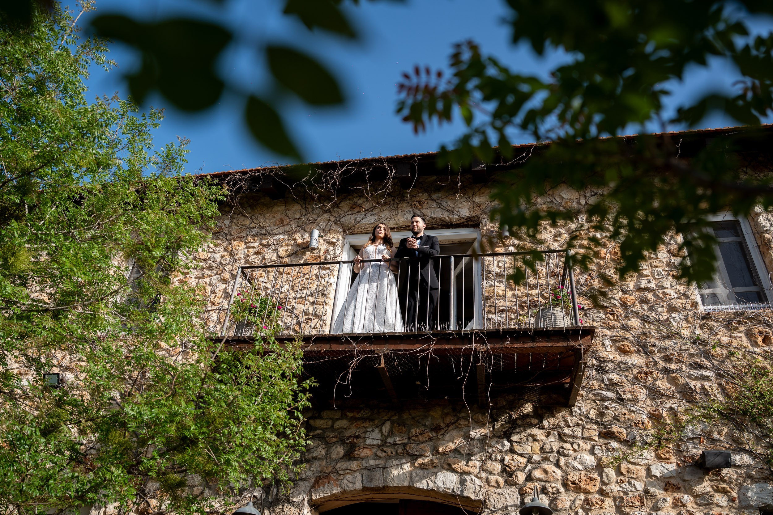 A bride and groom stand on the balcony of an old stone building surrounded by greenery