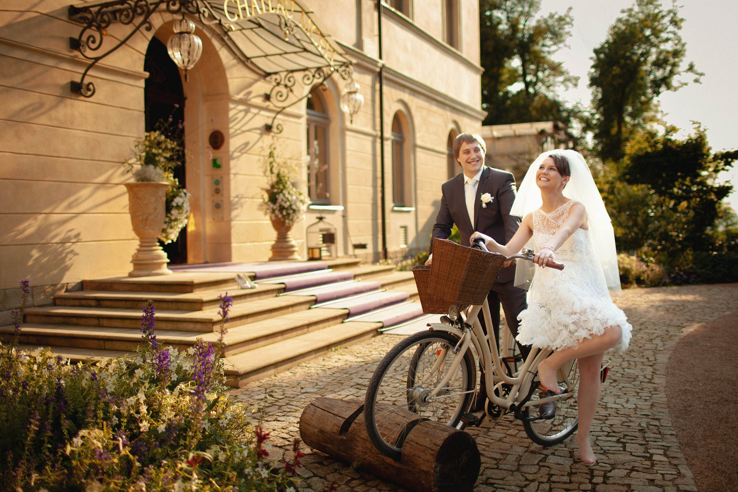 Happy newlyweds share a laugh as they stand near bicycles in front of the historic facade of the Chateau Mcely.