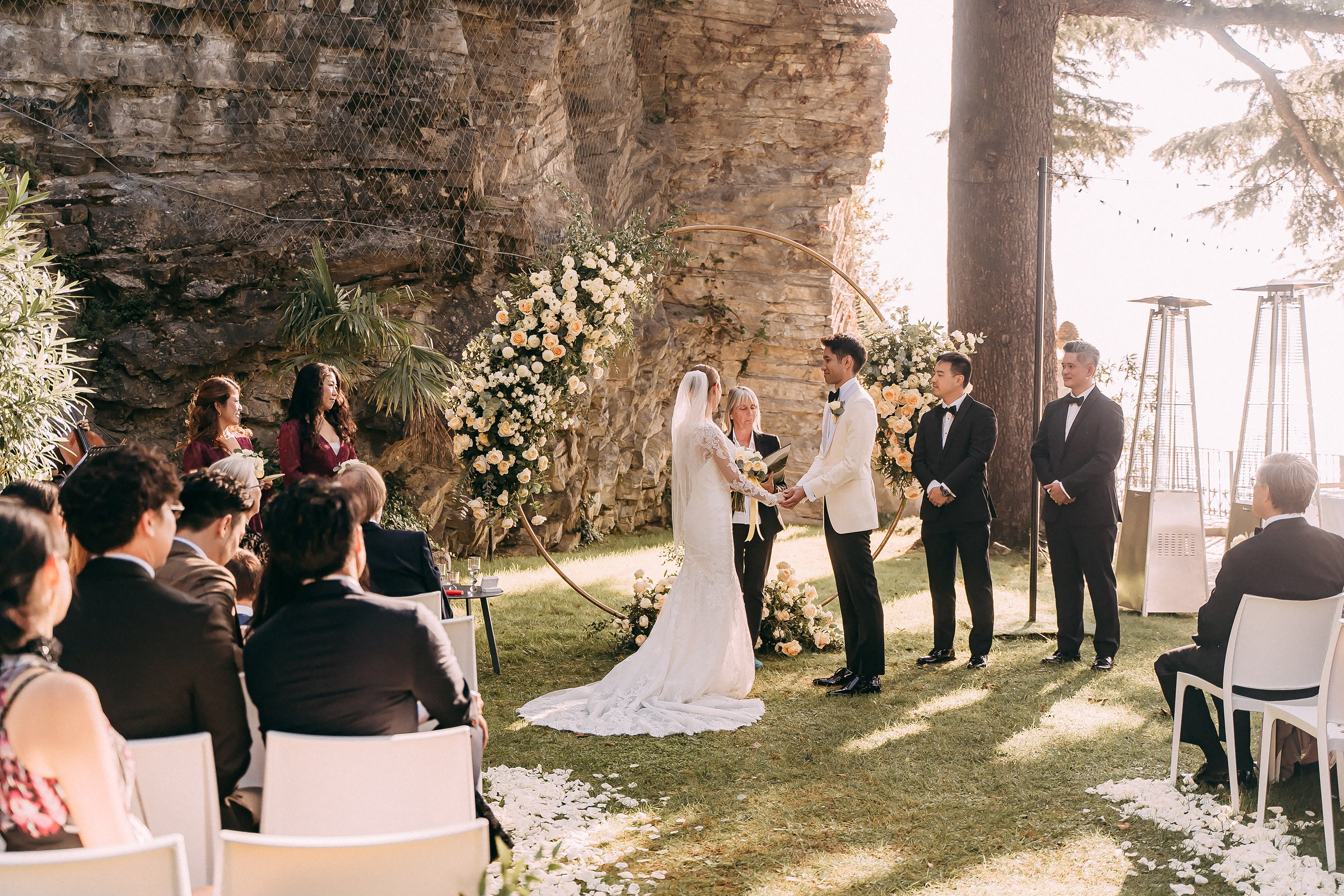 Bride and groom hold hands at the altar, surrounded by floral arrangements and guests seated nearby.