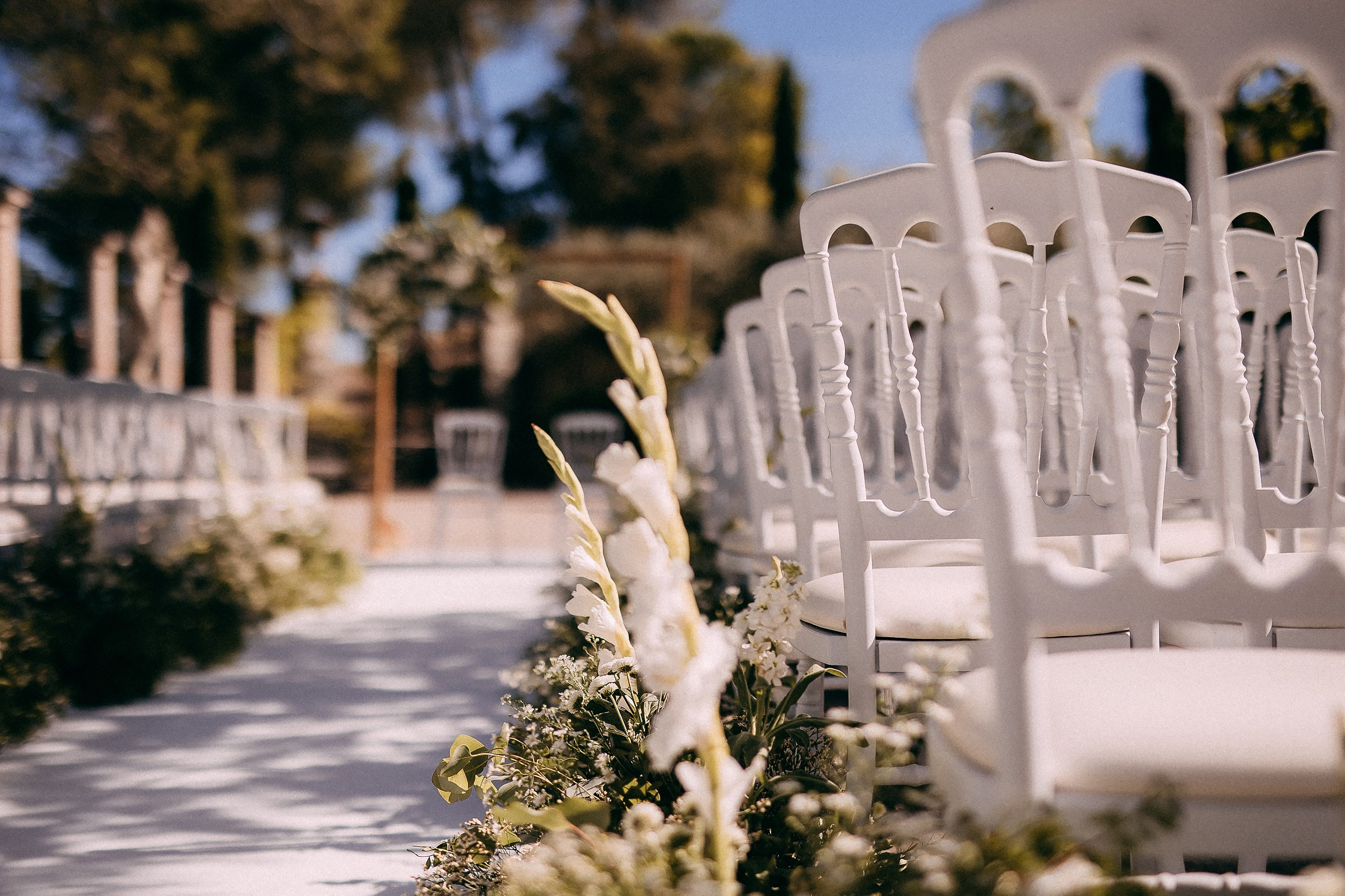Rows of elegant white chairs with floral decorations lining the aisle, set outdoors under a sunny blue sky in Provence.