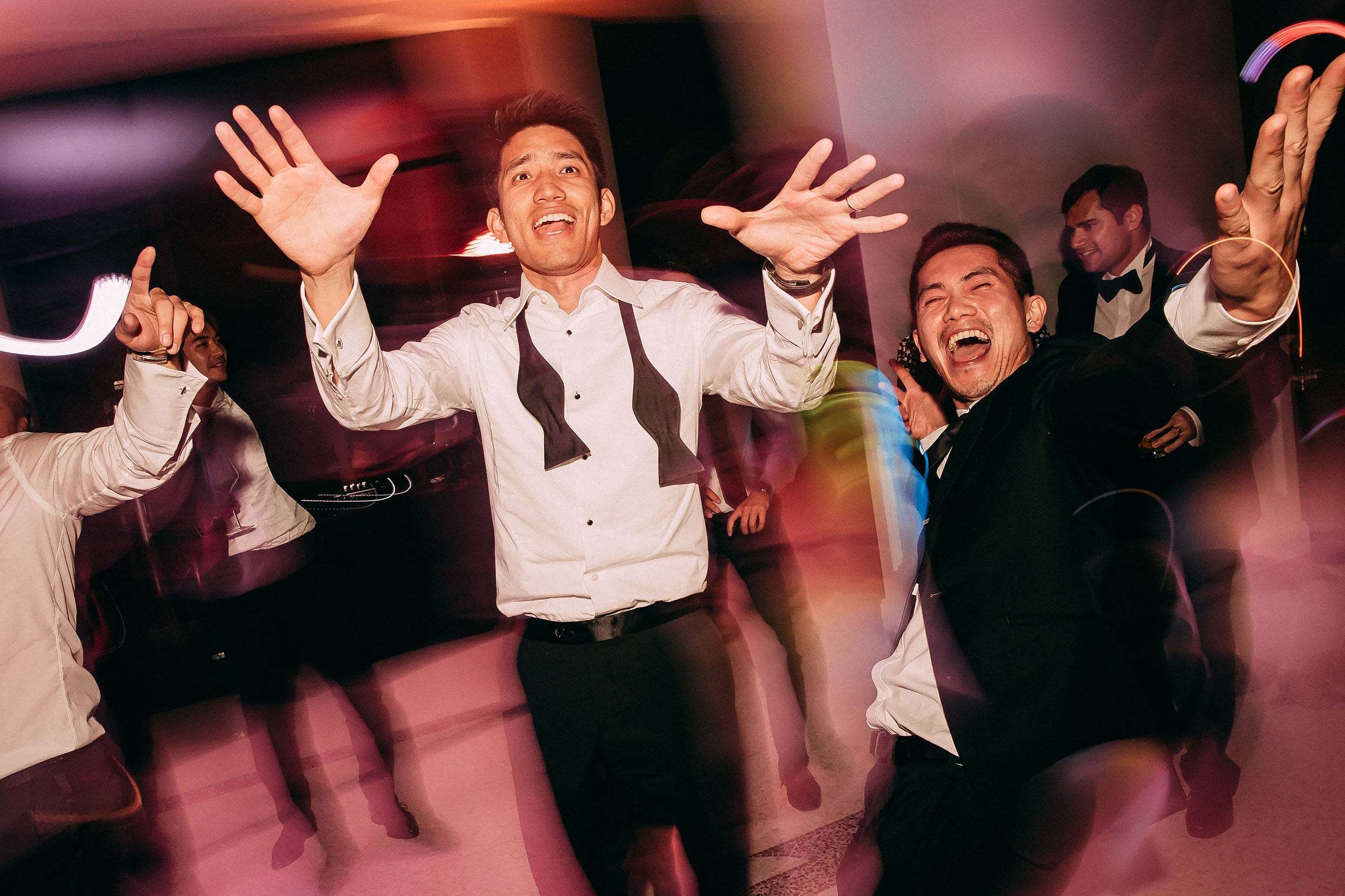 Partygoers, with two men in the foreground, dancing with their hands raised high, enjoying the vibrant reception.