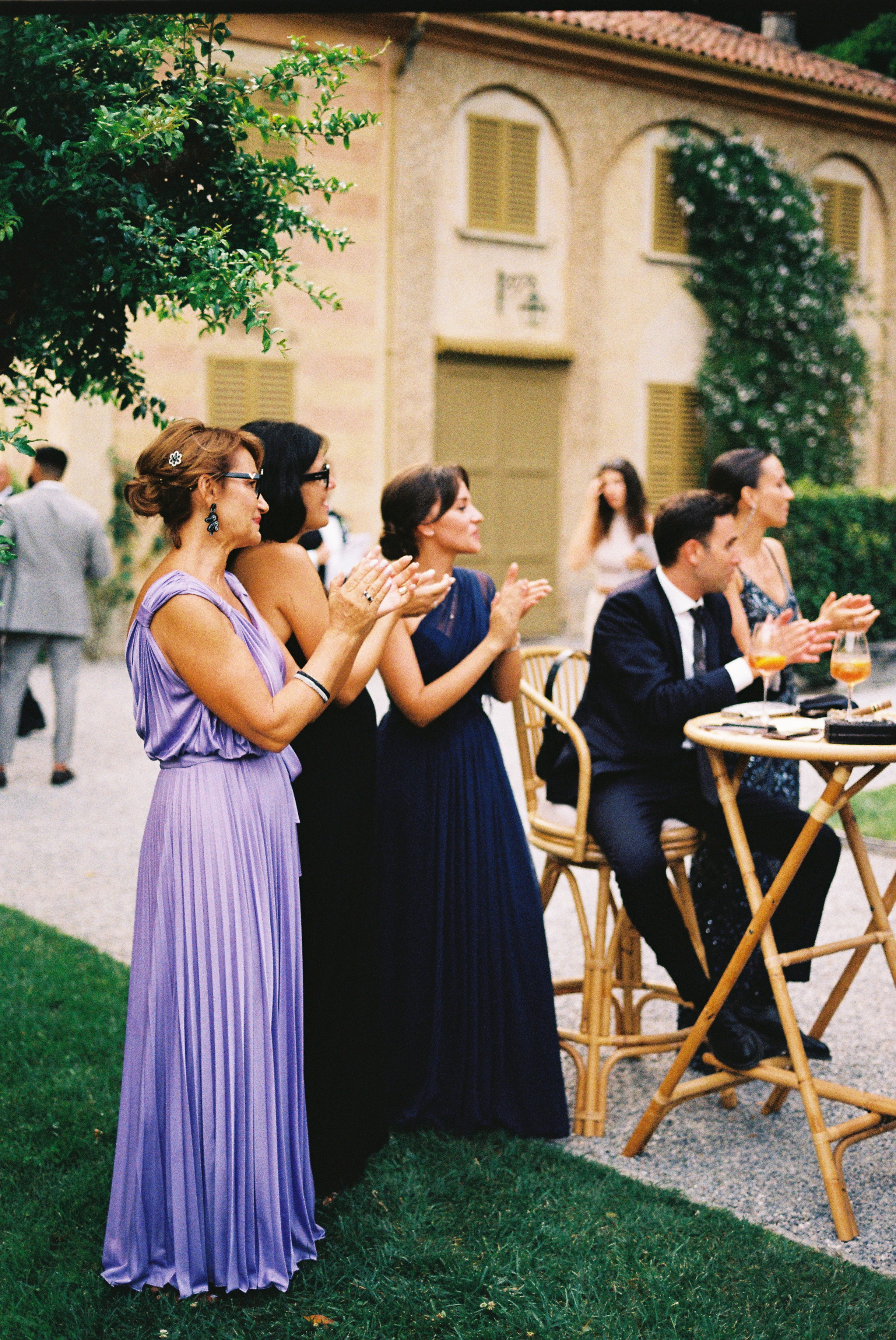 Women in elegant dresses applaud, standing near high-top tables at the reception.