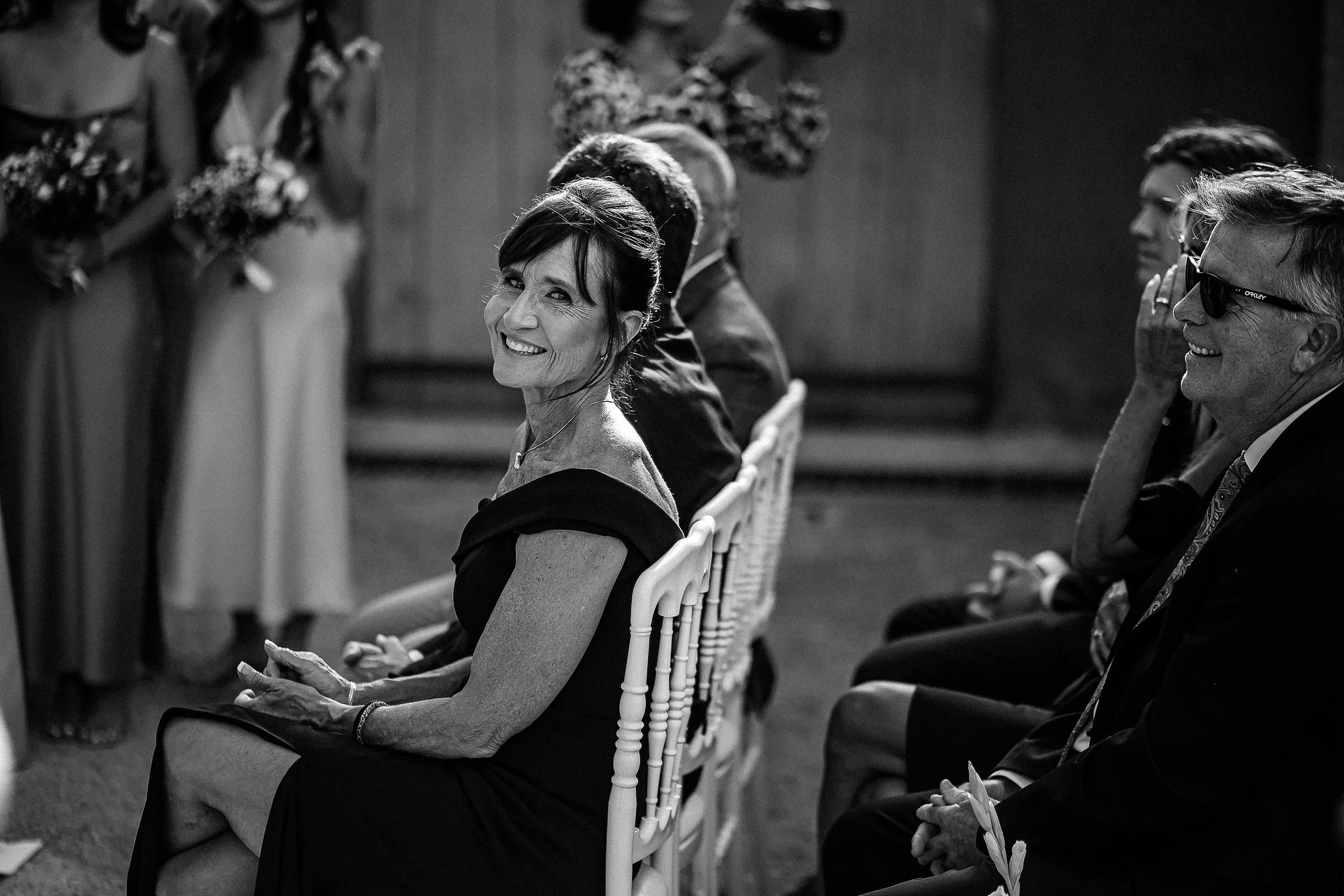A woman seated among the wedding guests turns back to smile warmly at the camera, surrounded by family and friends.