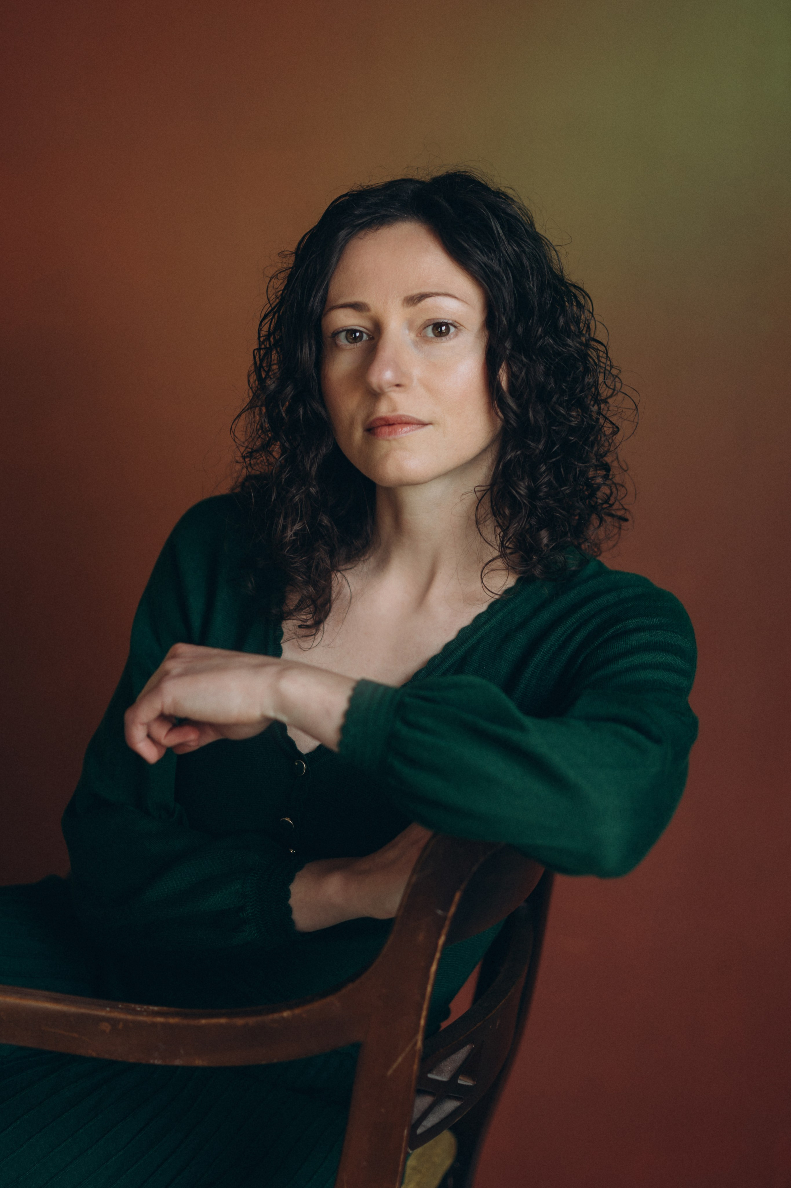 Actor headshot of a confident woman with curly hair in a green top, sitting on a chair against a brown background