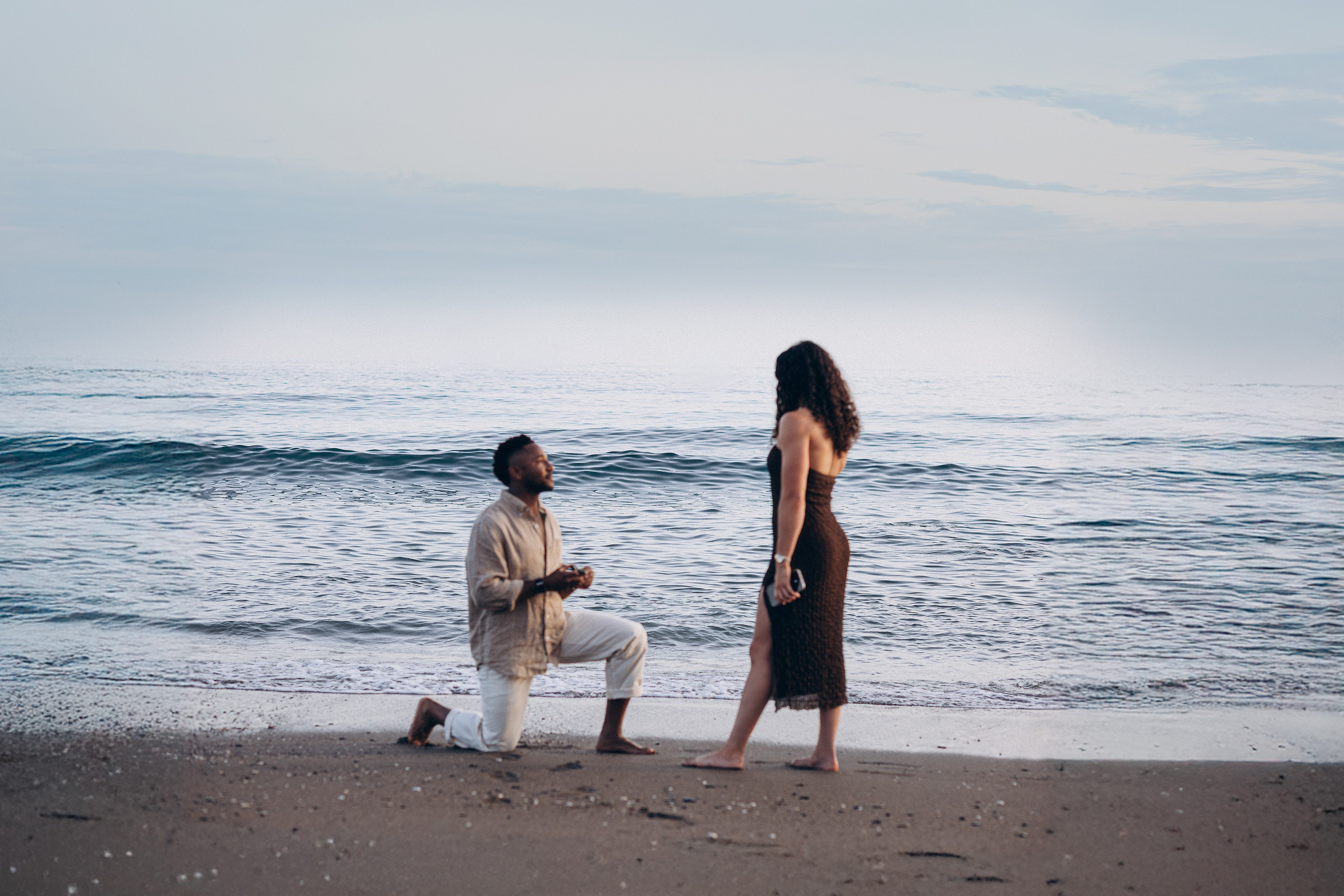 Romantic destination wedding proposal on a beach in Málaga, Spain, with groom kneeling on the sand as Mediterranean waves roll behind the couple at sunset.