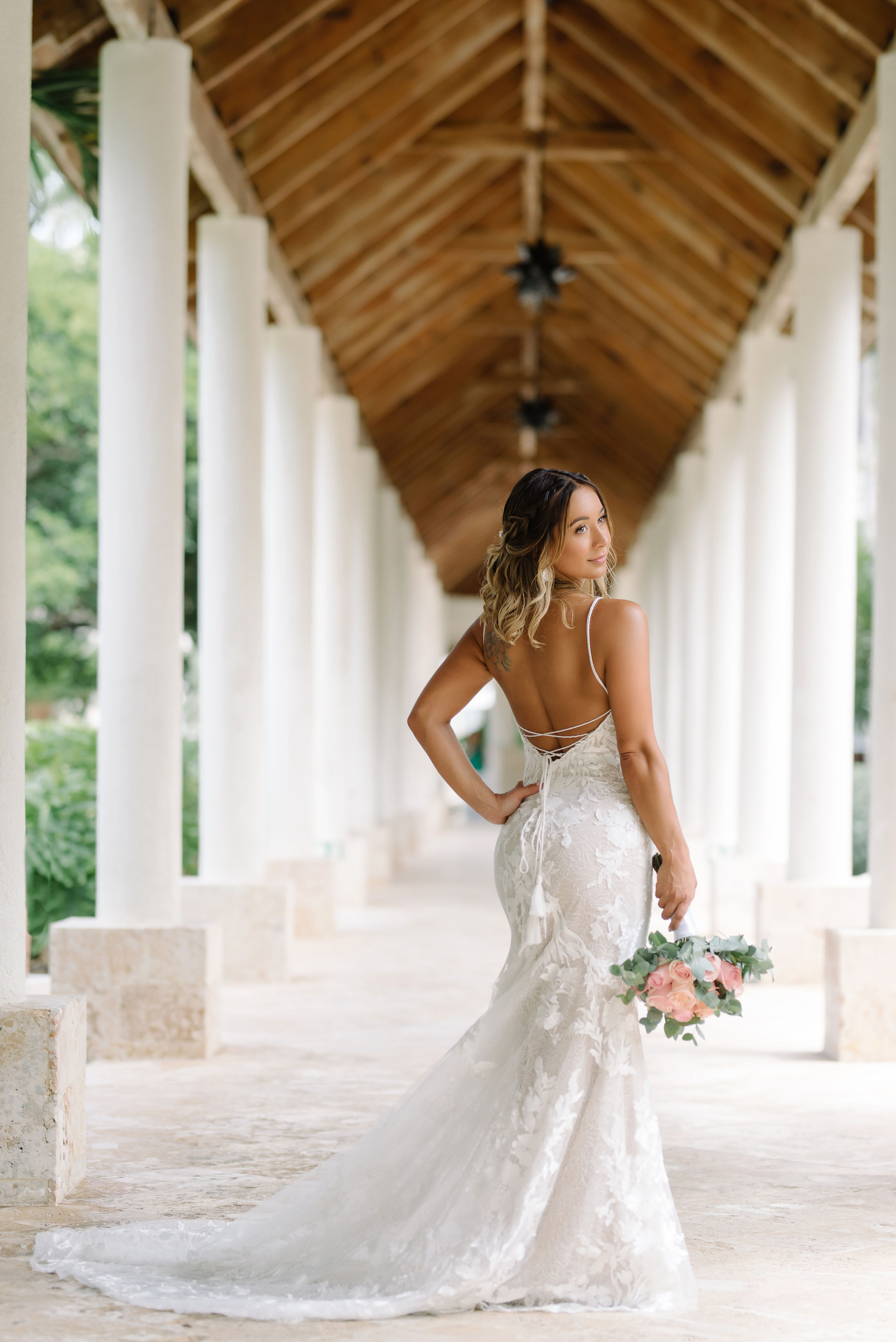 Beautiful bride in a lace wedding dress holding a bouquet of pink and peach roses, standing in an elegant columned hallway with a wooden ceiling