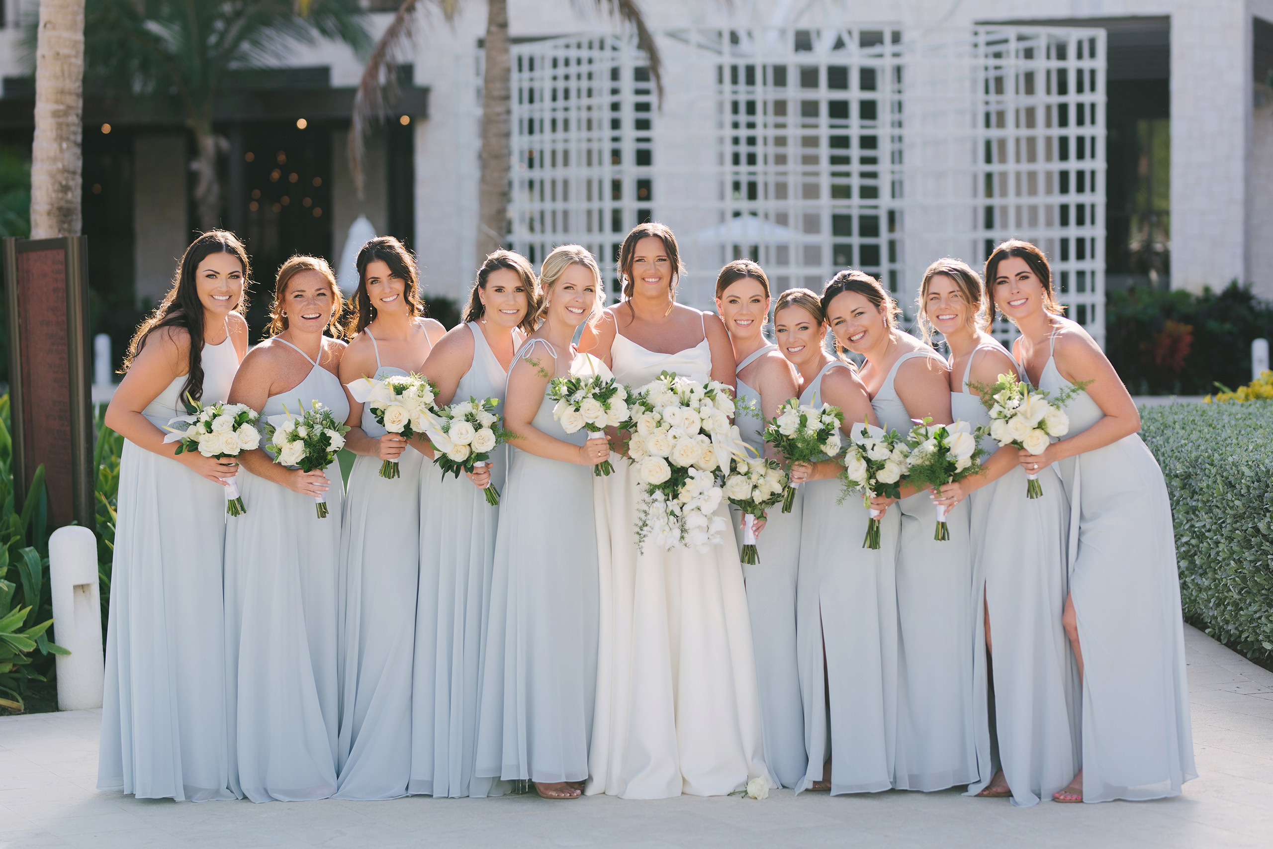 Bride with her bridesmaids wearing light blue dresses, holding white and green floral bouquets, posing outdoors in a sunny, elegant setting