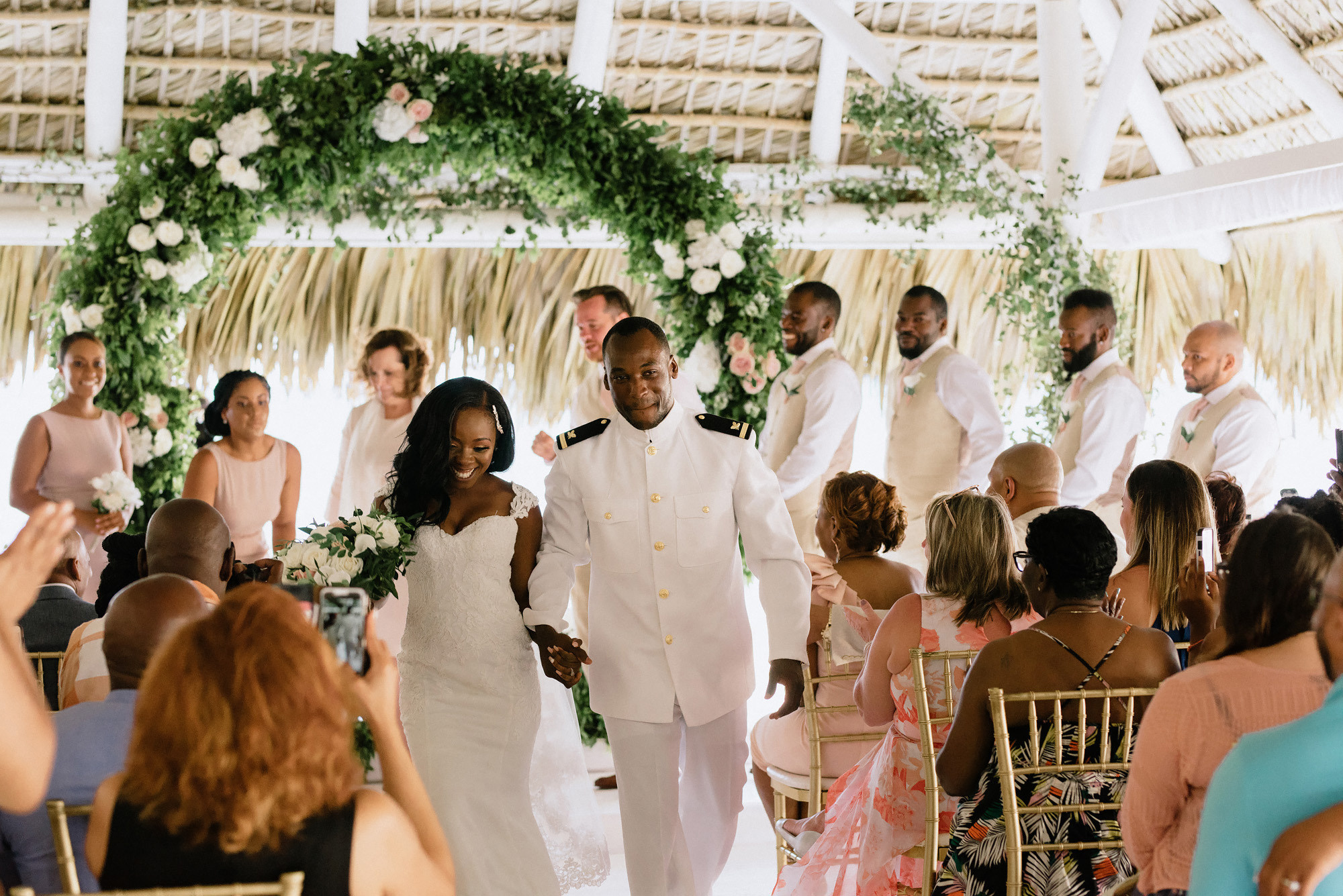Smiling bride and groom walking down the aisle under a lush floral arch during their tropical wedding ceremony, surrounded by guests and bridal party