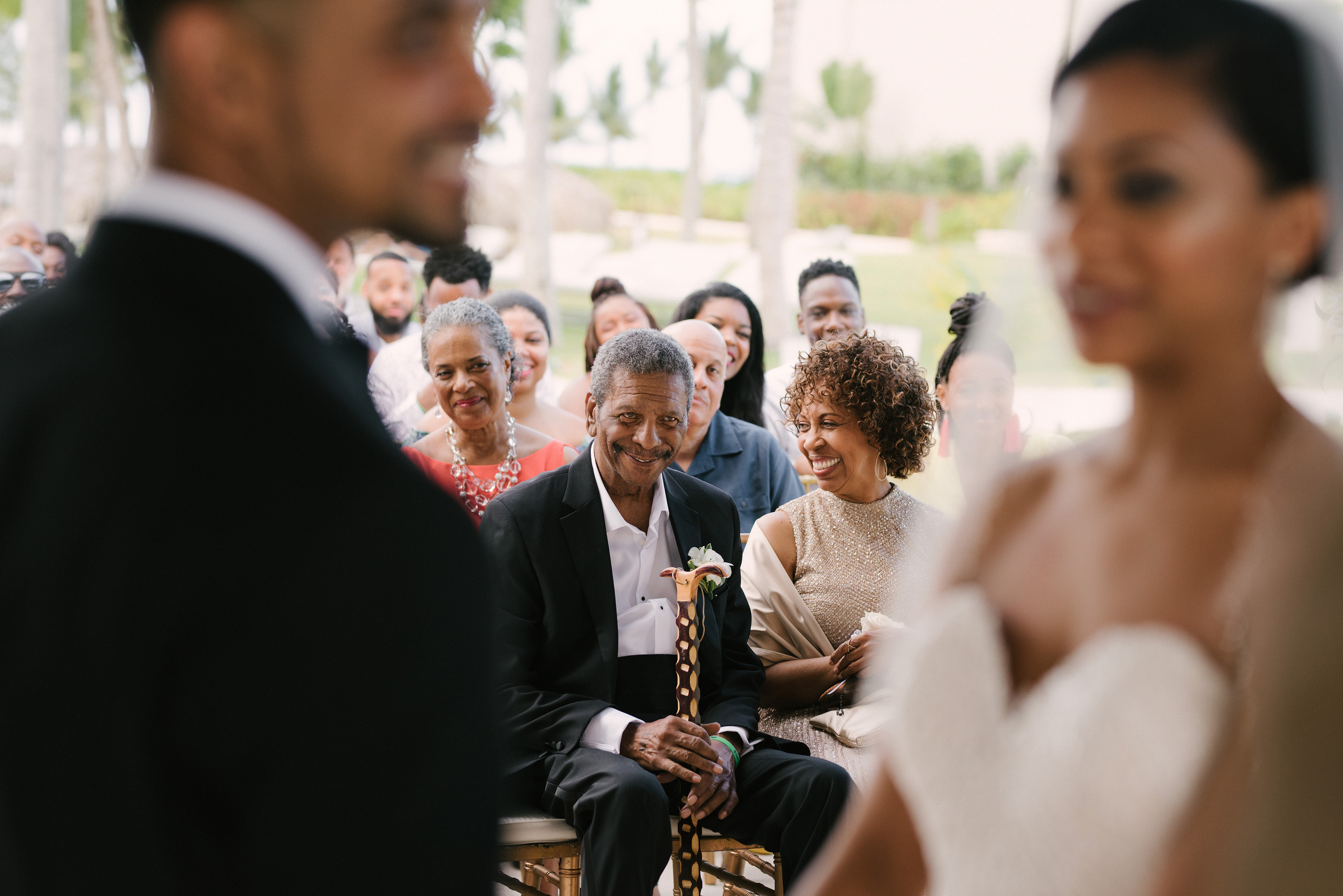 A heartfelt moment during a wedding ceremony as the guests, including an elderly man with a cane and a woman in an elegant gown, smile with joy and pride. The focus on their expressions captures the emotional connection and happiness shared by loved ones witnessing the union. The blurred foreground of the bride and groom adds depth to this intimate scene