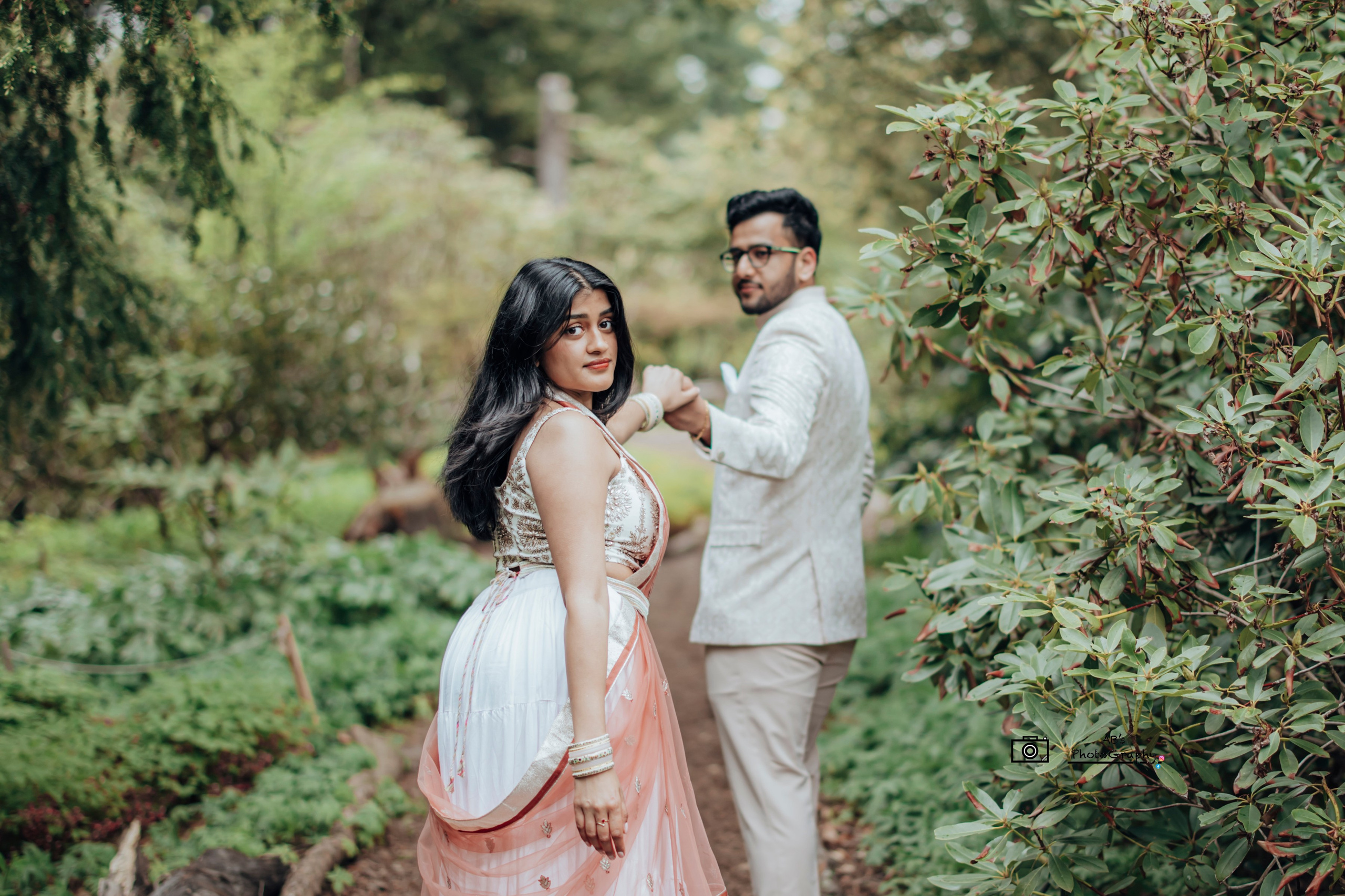 Romantic outdoor pre-wedding portrait of an Indian couple holding hands on a garden pathway, dressed in elegant traditional outfits, captured in soft natural light by Abhishek B Photography.