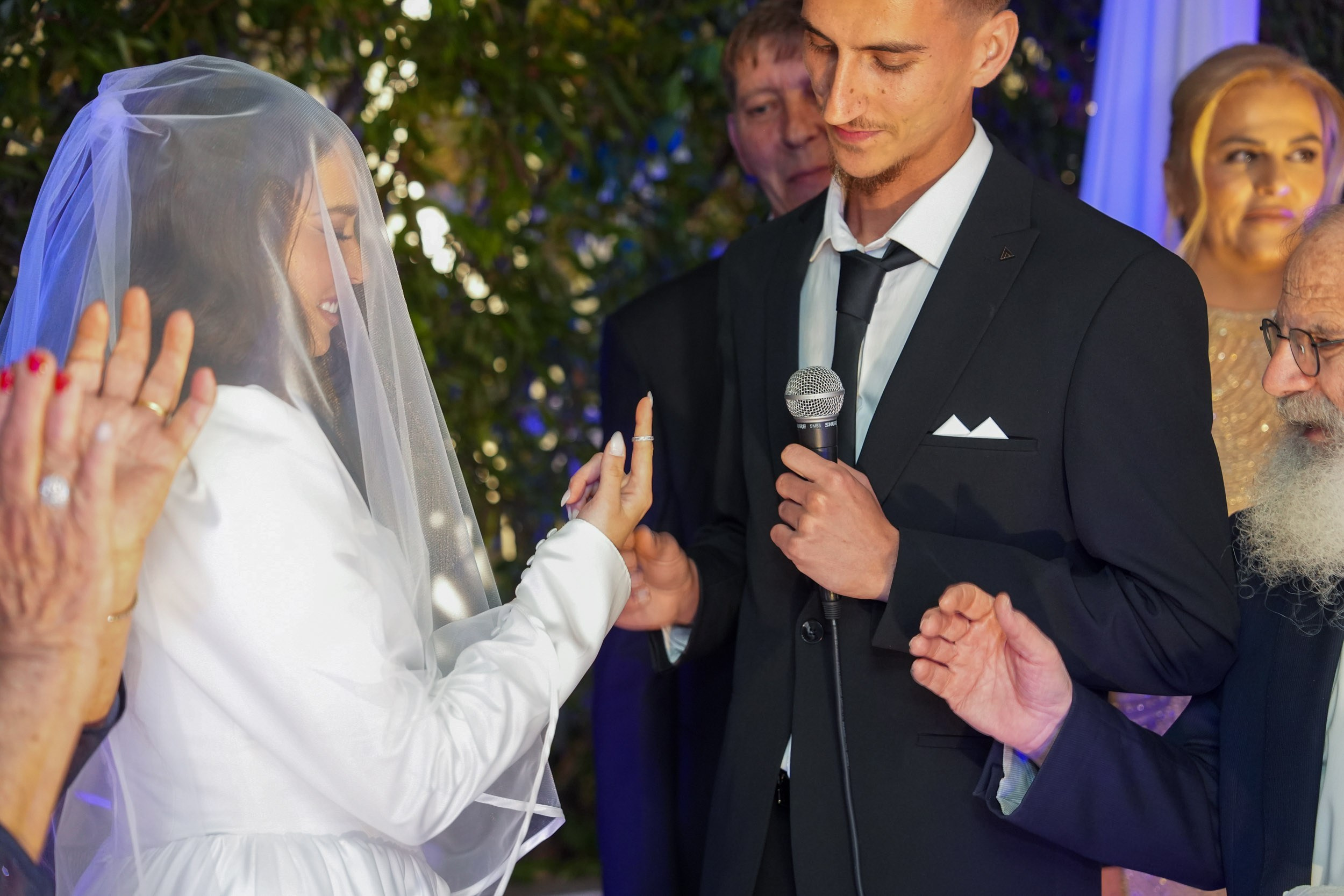 Groom Garik speaking into a microphone while placing a ring on bride Lin's finger under the Chuppah at Kadita Event Garden in Emek Hefer, Israel